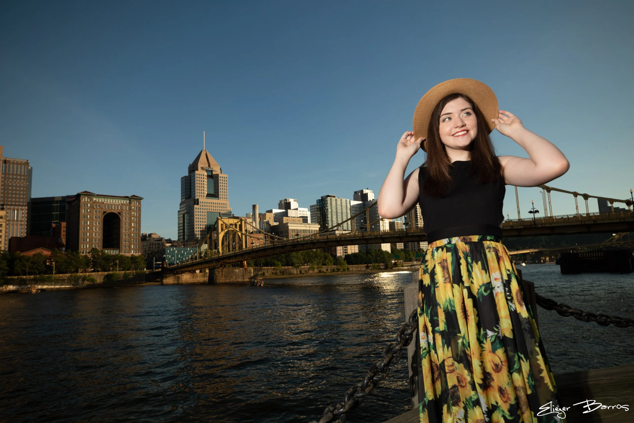 A woman wearing a straw hat and a black top with a colorful skirt stands on a bridge by the river, smiling, with city buildings and a bridge in the background during sunset.
