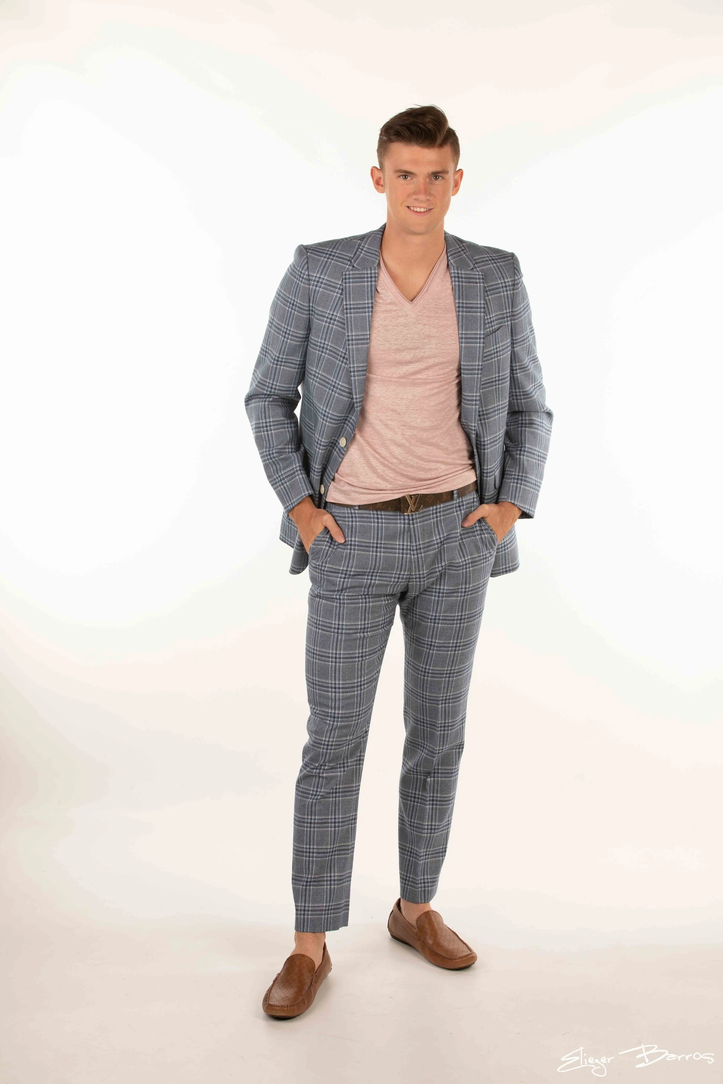 A young man in a gray plaid suit with matching pants and blazer, pink V-neck T-shirt, brown flats, and a black belt with a gold logo, standing with hands in pockets against a white background, smiling at the camera.