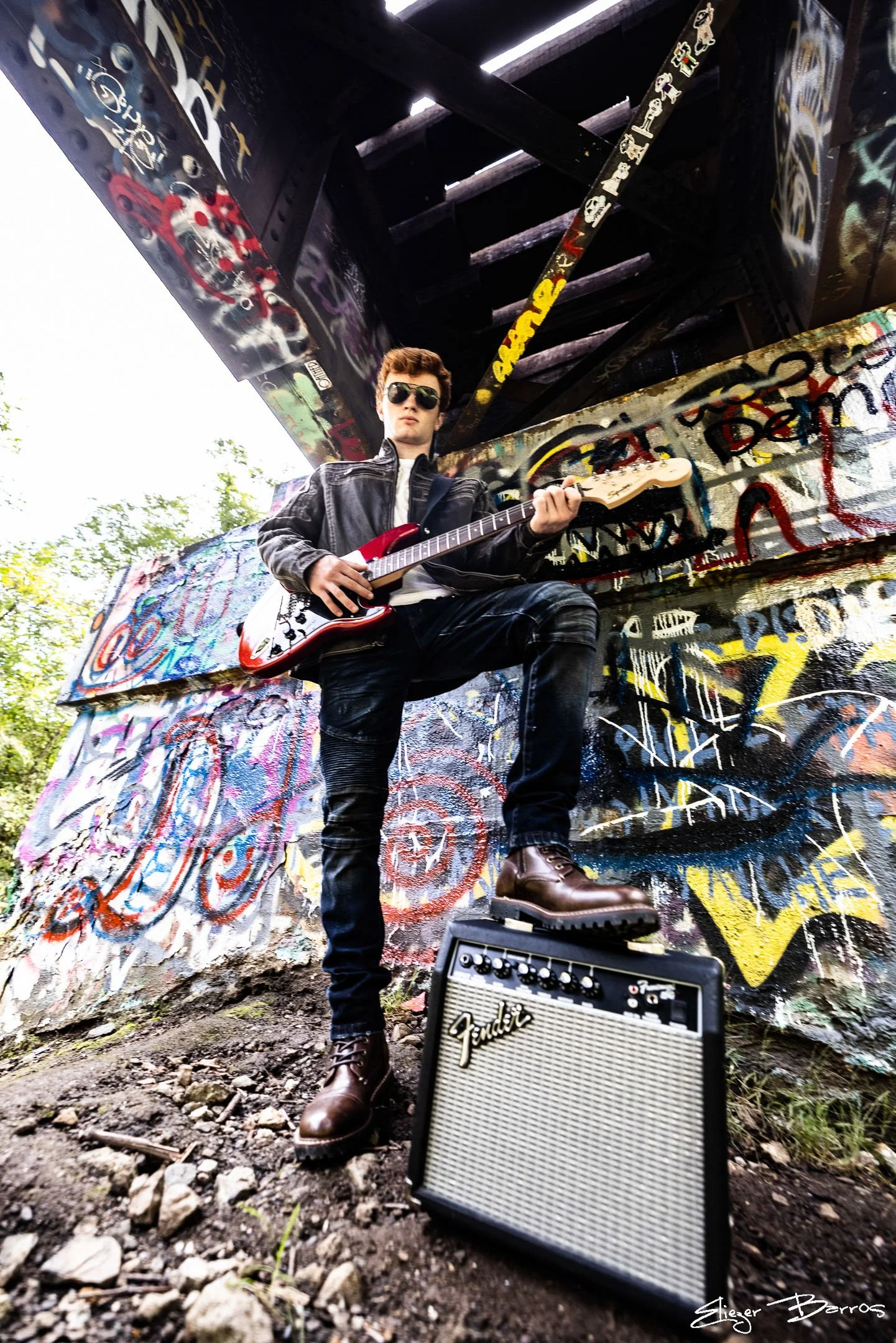 Young man with sunglasses playing electric guitar under a graffiti-covered bridge with a Fender amp in the foreground.