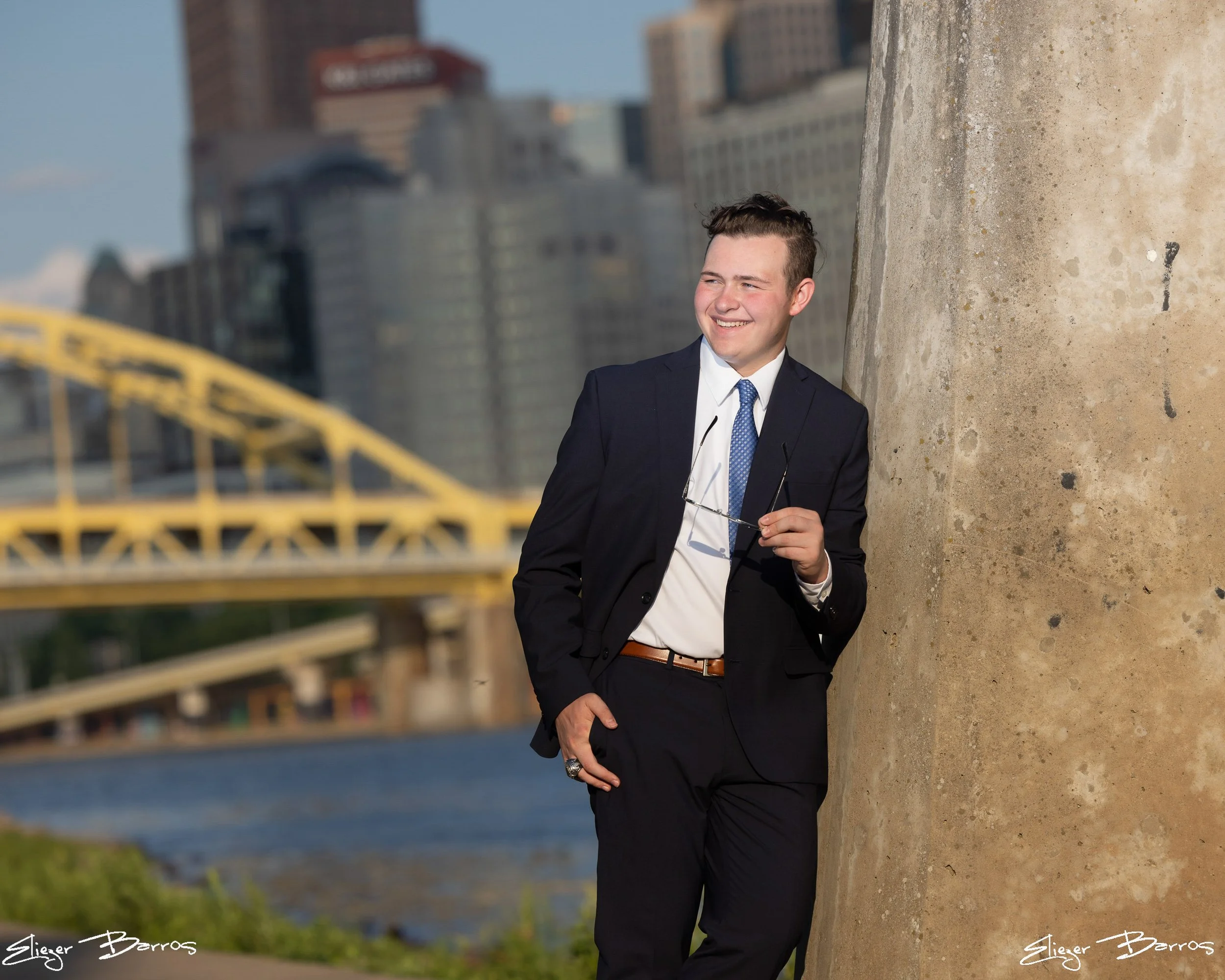 A young man in a dark suit and tie holding glasses, smiling while leaning against a large concrete structure outdoors with a cityscape and a yellow bridge in the background.
