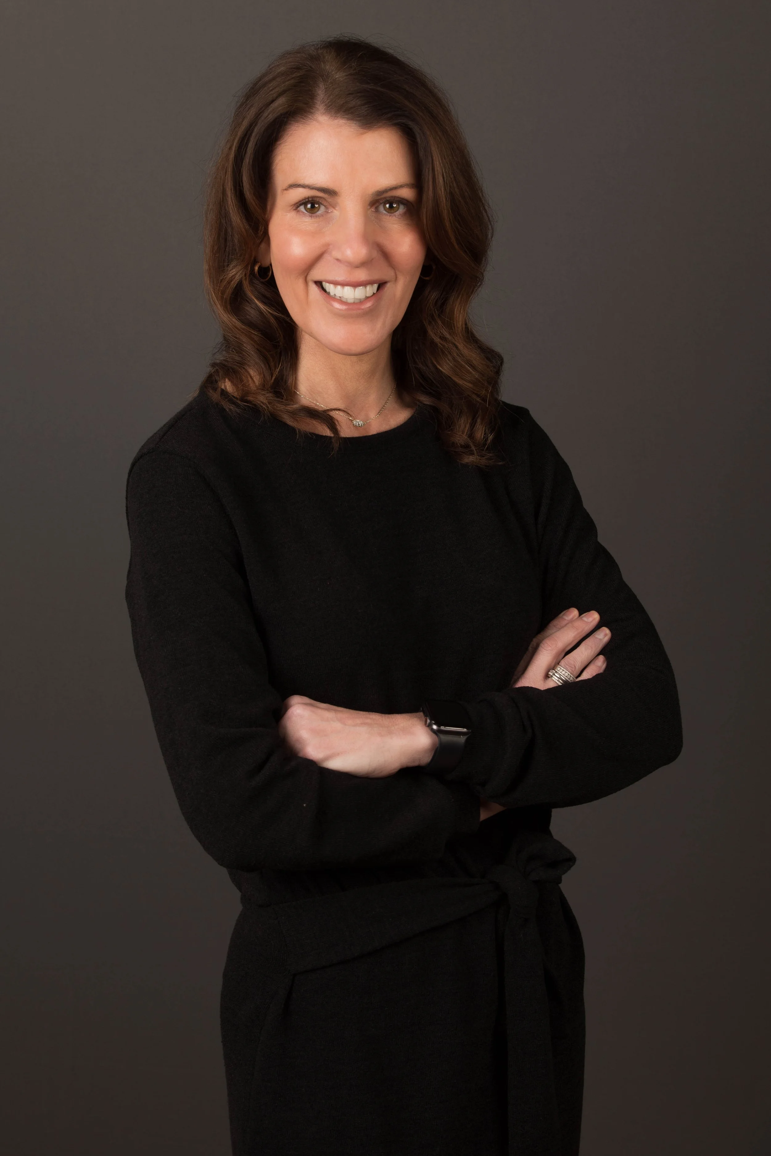 A woman with brown, shoulder-length, wavy hair wearing a black long-sleeve top, jewelry, and a smartwatch, standing against a dark gray background, smiling with arms crossed.