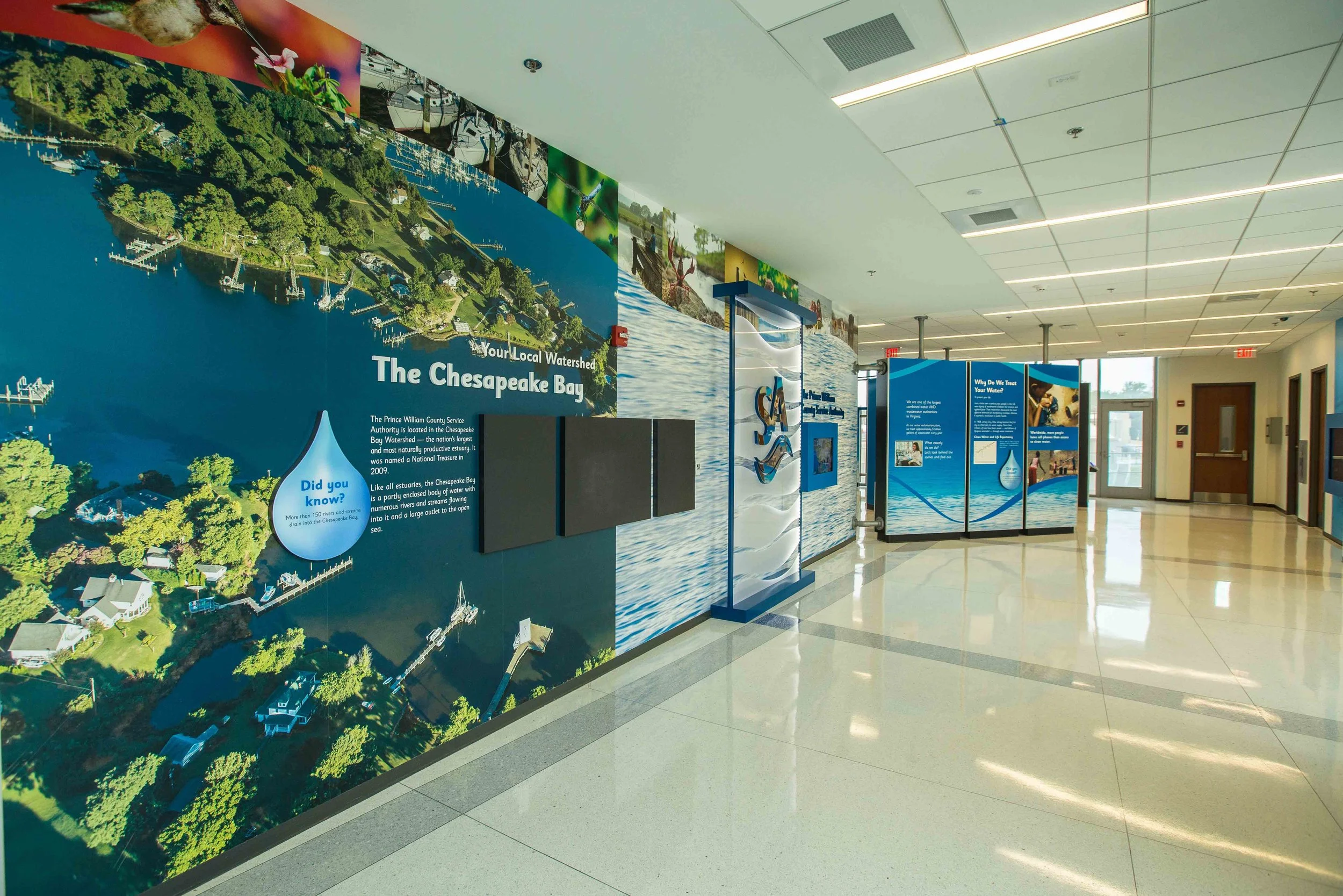 Indoor informational exhibit about Chesapeake Bay, featuring large aerial map and panels with water conservation information. The exhibit is located in a building hallway with tiled flooring, ceiling lights, and a glass door in the background.