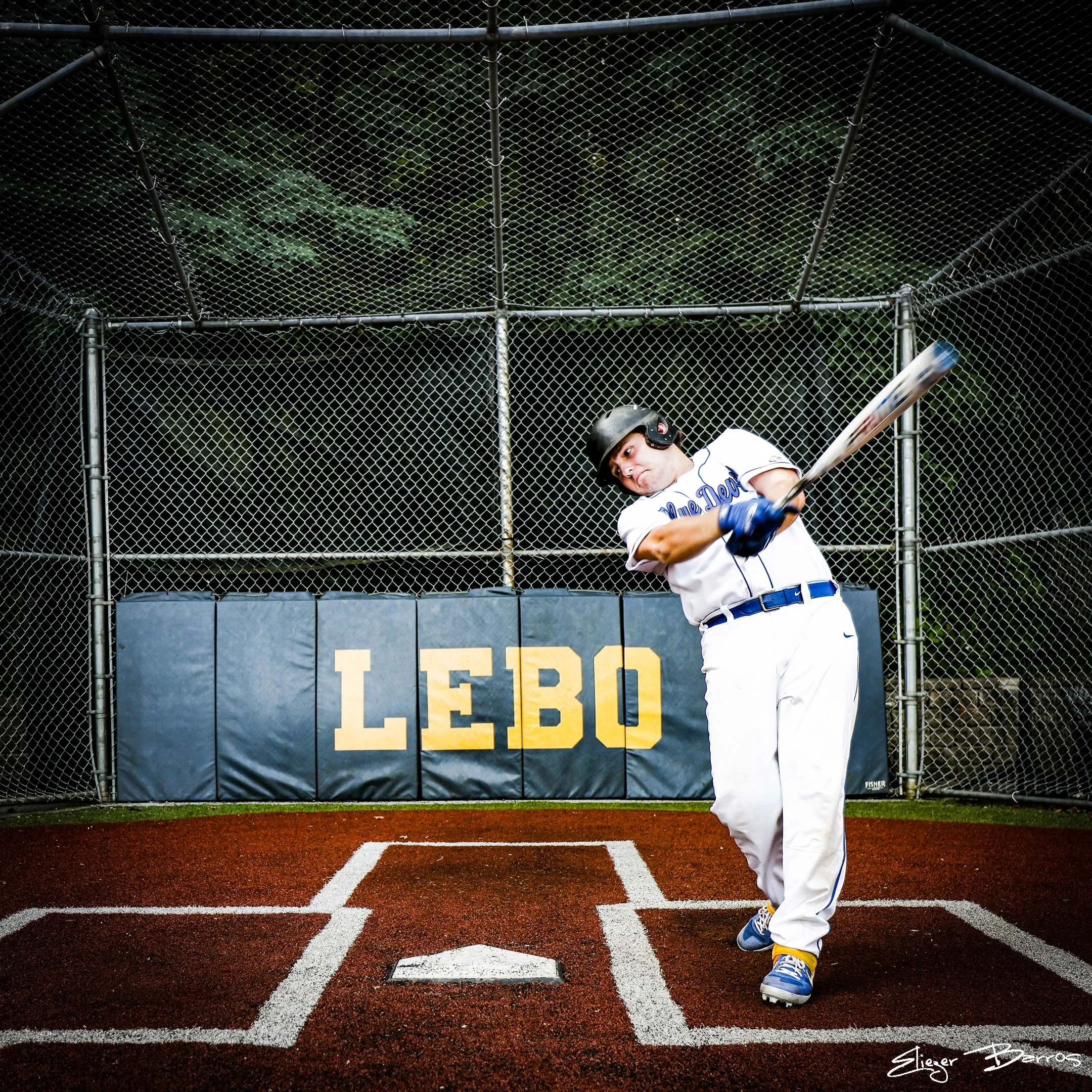A baseball player standing at home plate on a field, preparing to bat, with a chain-link fence behind him and a blue sign that says 'LEBO' in the background.
