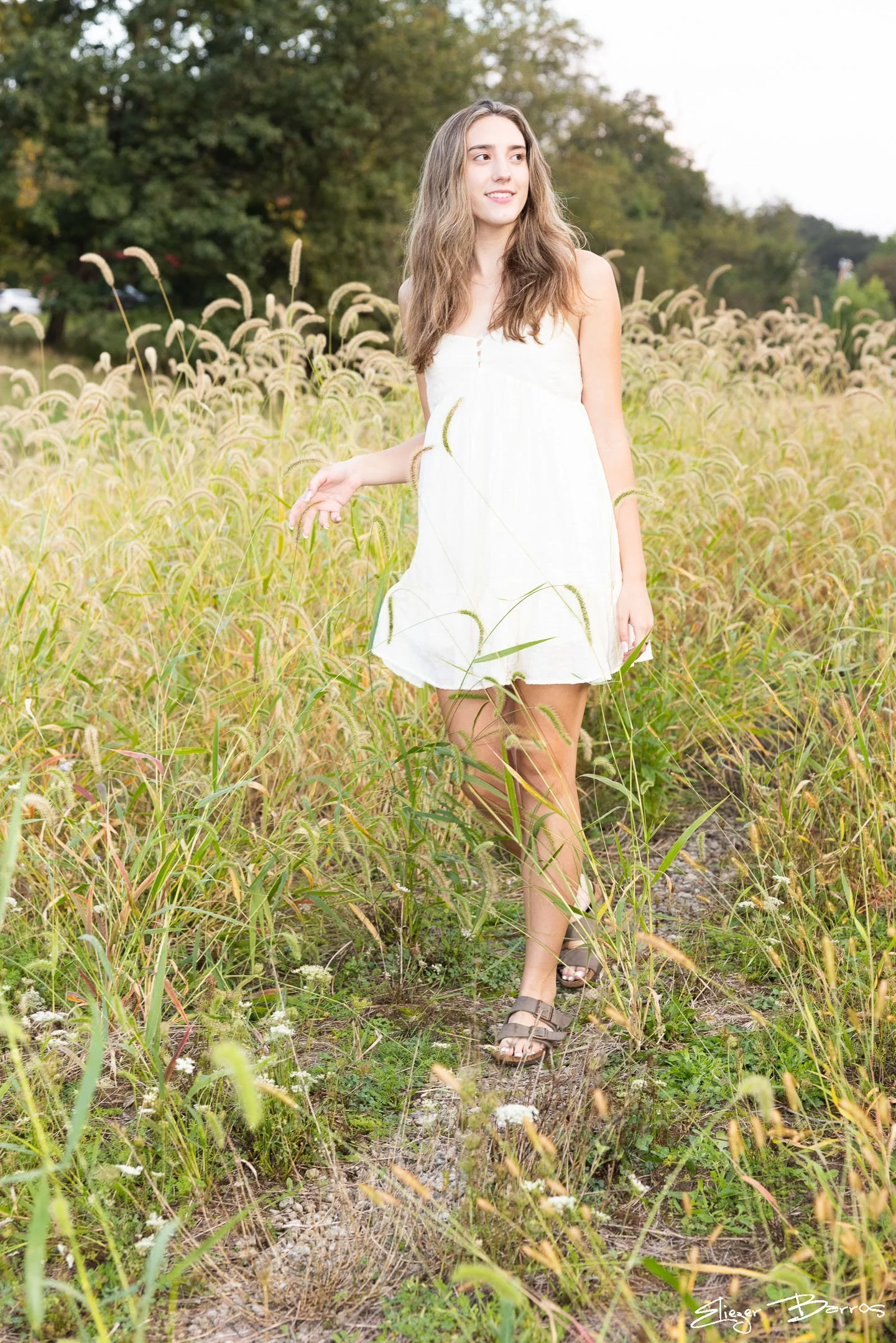 Young woman in a white summer dress walking through a field of tall grass and wildflowers, with trees in the background.