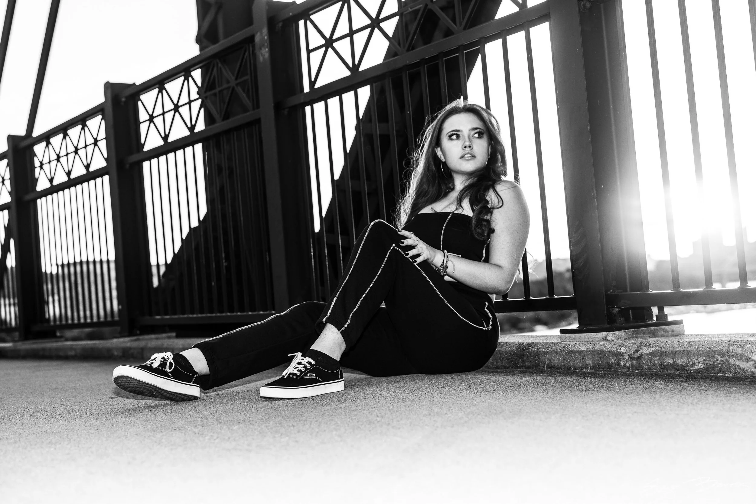 A young woman with long hair, wearing a black strapless top, black pants with white stripes, and sneakers, sitting on the ground with her back against a railing on an urban rooftop, with city buildings and the sky in the background.
