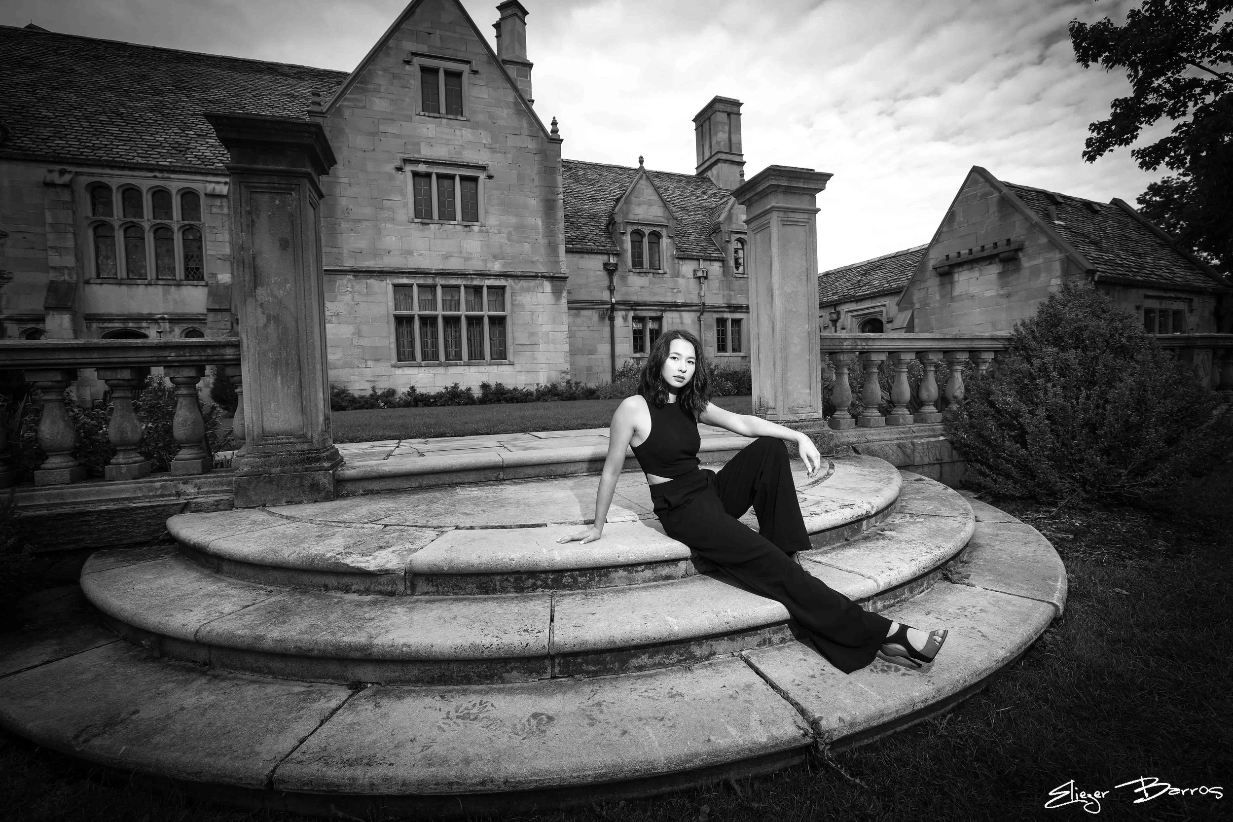 A young woman with shoulder-length hair sitting on a circular stone platform in front of a large, historic stone building with multiple chimneys and windows, surrounded by a manicured lawn and bushes. The image is in black and white.