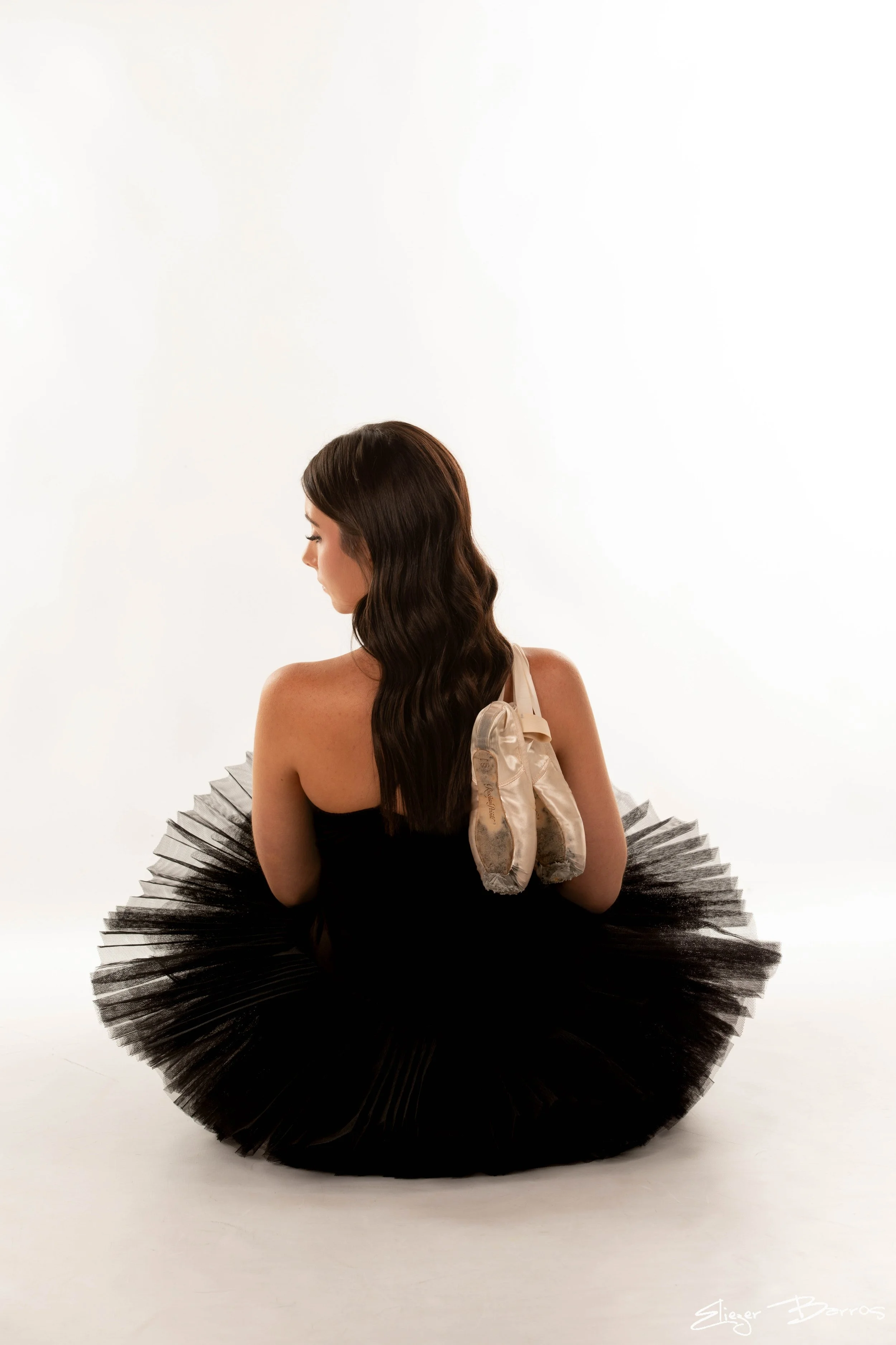 A young woman with dark brown hair in loose waves, wearing a black strapless dress and a large black tutu, is sitting with her back turned to the camera against a plain white background.