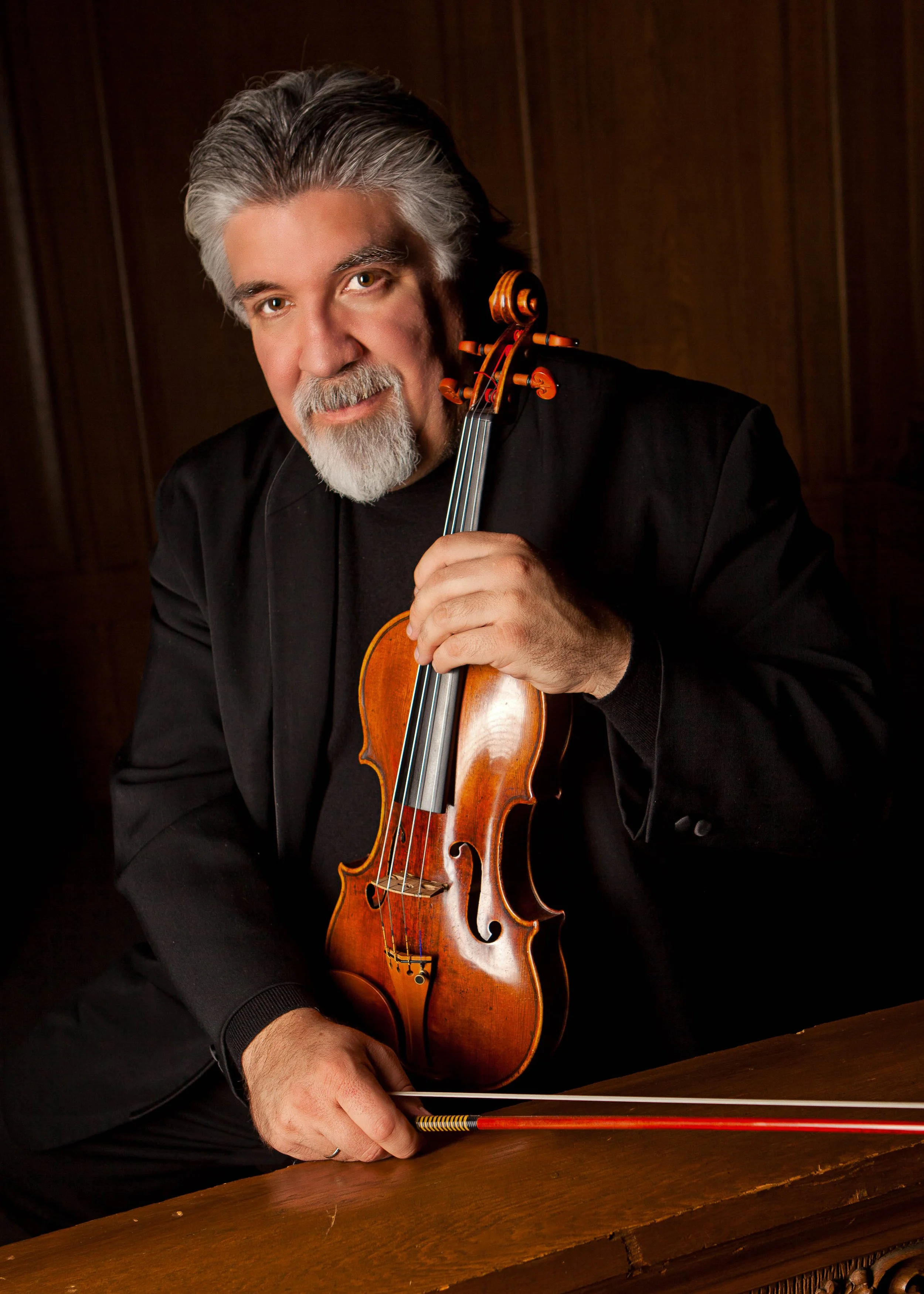 A man with gray hair and beard holding a violin and bow, sitting at a wooden table against a dark wooden wall.