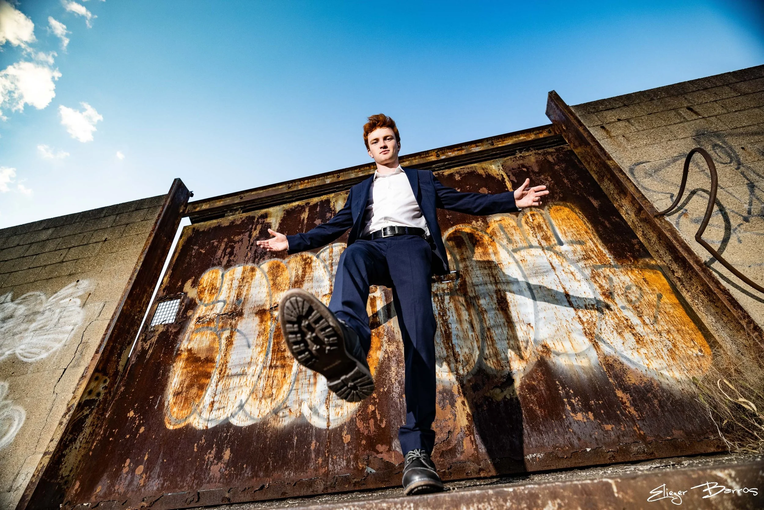A young man in a suit standing on a rooftop amidst graffiti and rusted metal, with a clear blue sky in the background. He is looking at the camera with his arms outstretched and one foot slightly raised.