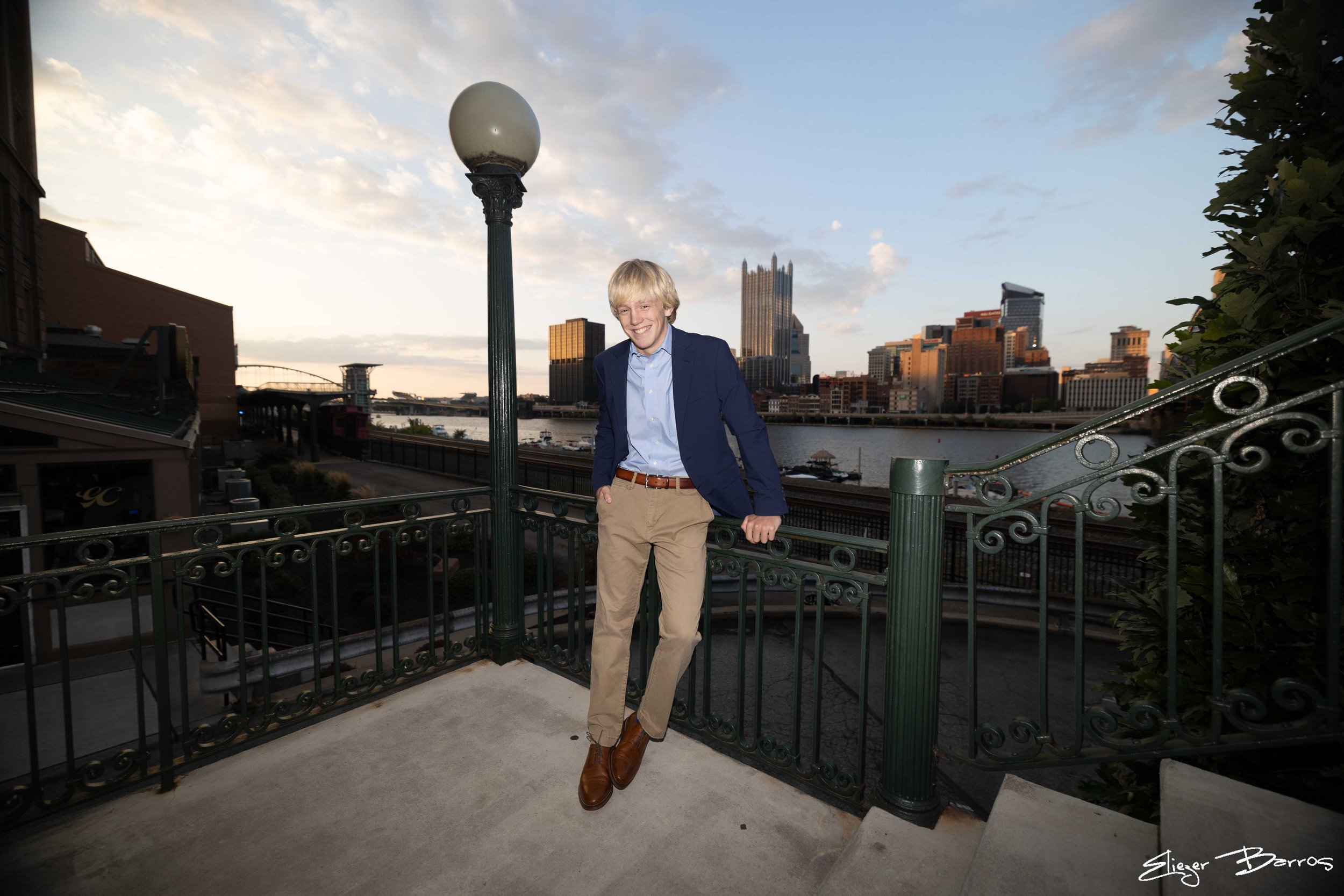 A smiling young man with blond hair wearing a navy blazer, light blue shirt, khaki pants, and brown shoes, standing on a balcony with a city skyline and river in the background during the evening.