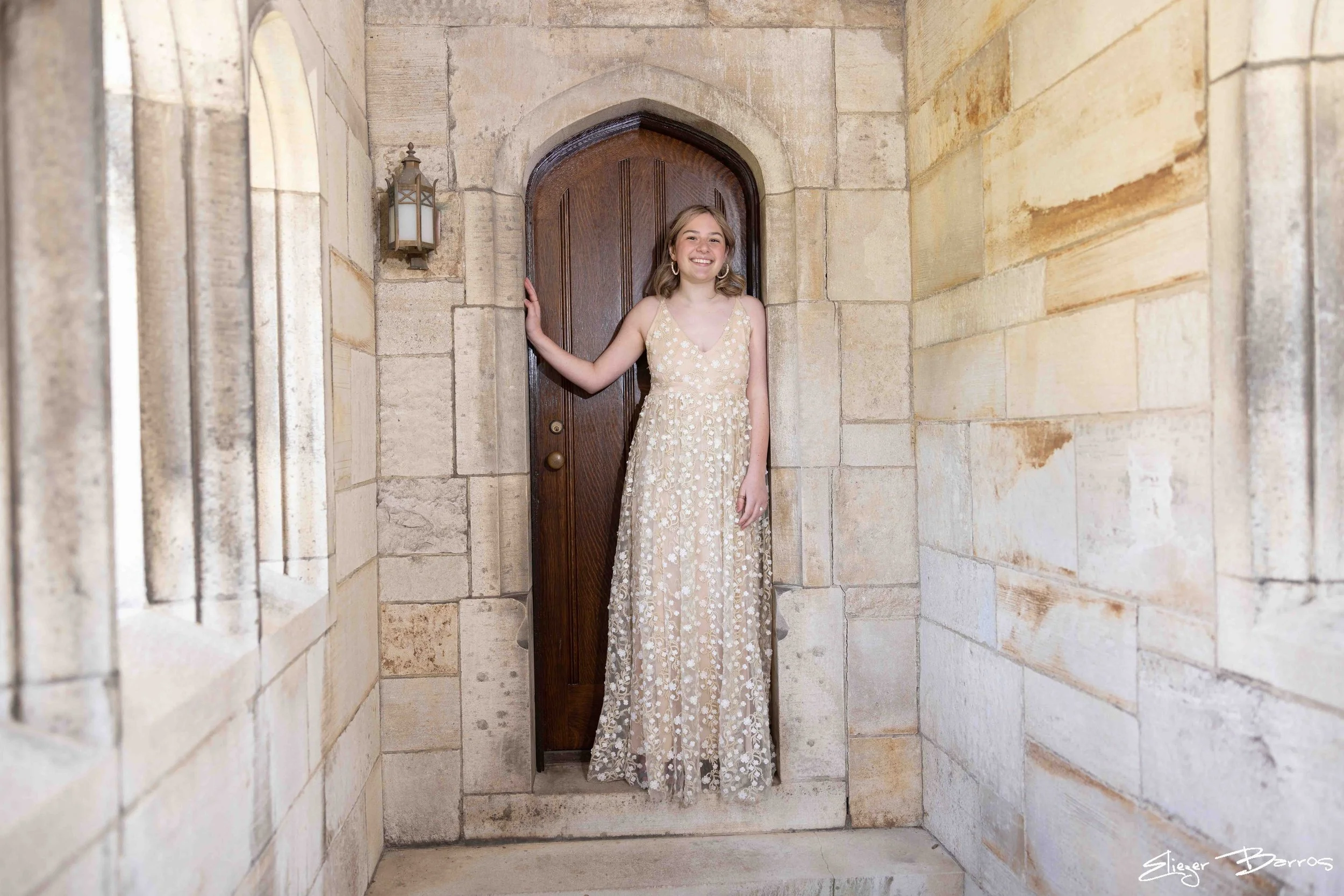 A young woman in a long, elegant, cream-colored dress with floral patterns, standing in a doorway with stone walls and a wooden door. She is smiling and has shoulder-length blonde hair.