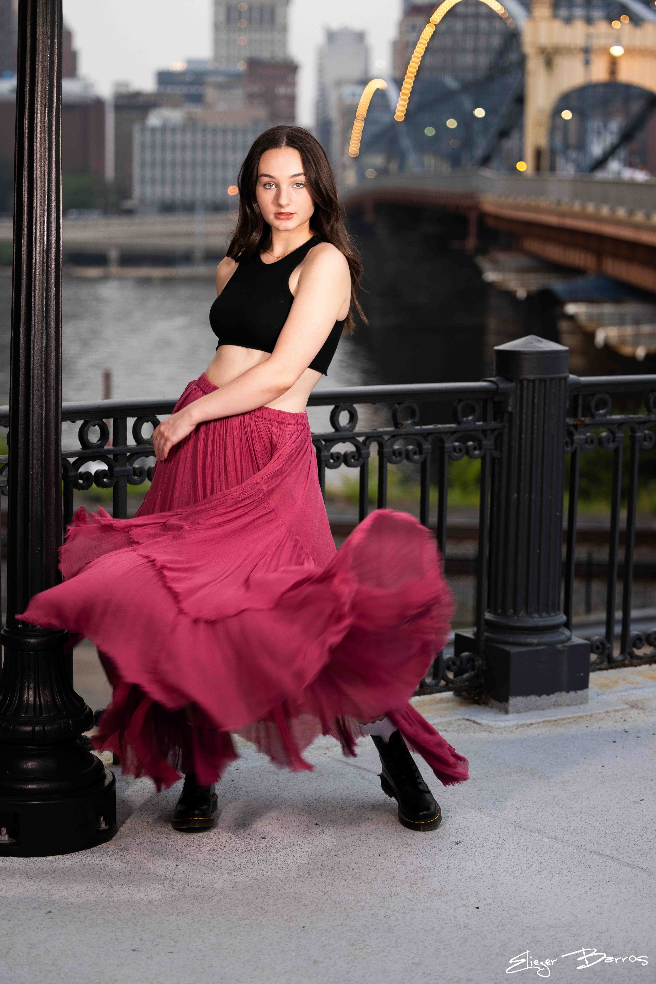 A young woman with long dark hair, wearing a black crop top and a flowing pink skirt, stands by a black metal fence along a river in an urban setting during evening. The city skyline with tall buildings is blurred in the background, and the woman is 