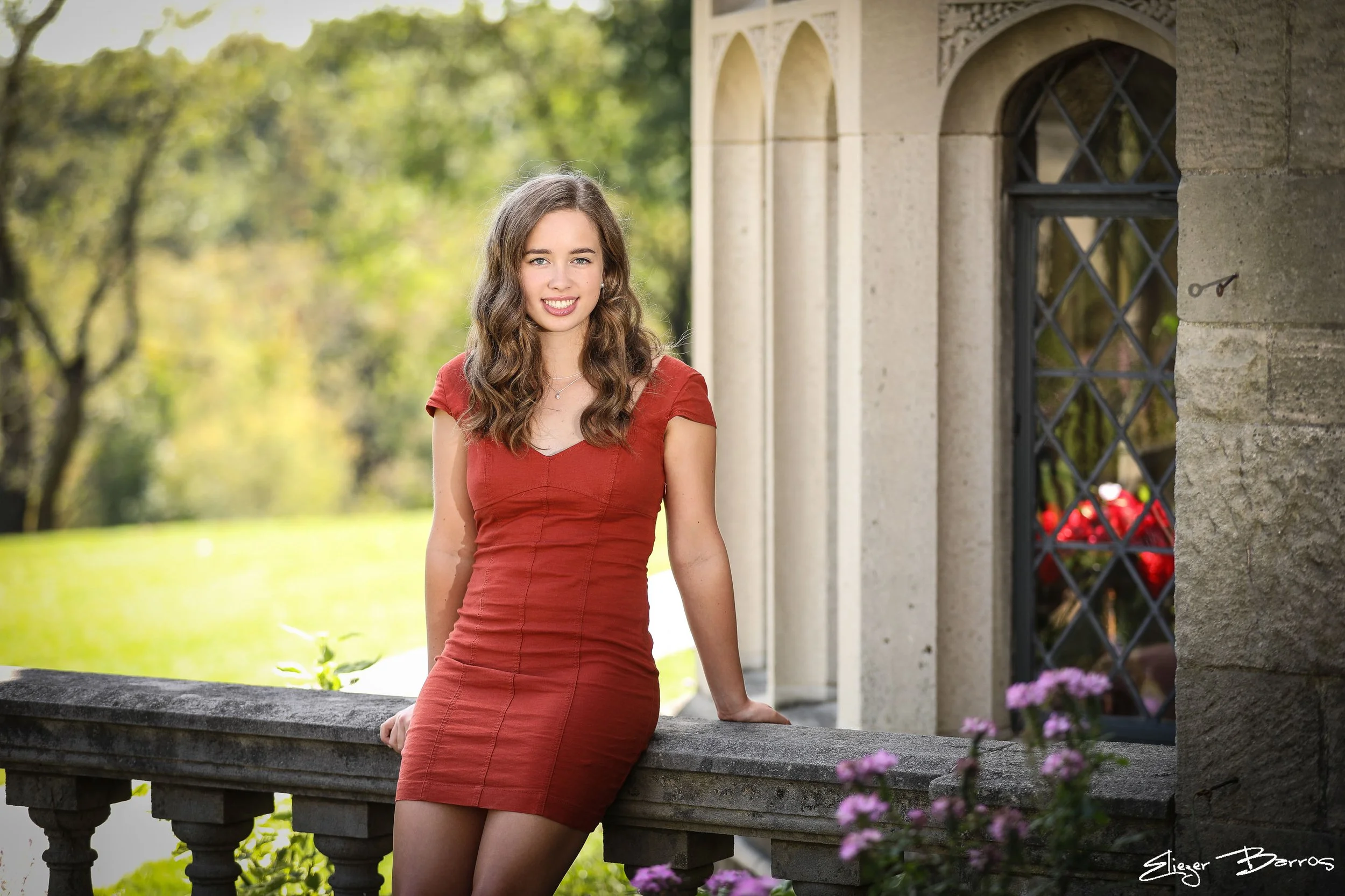 A young woman with long, curly brown hair and wearing a red dress, leaning against a stone ledge outside near a historical building with arched windows and greenery in the background.