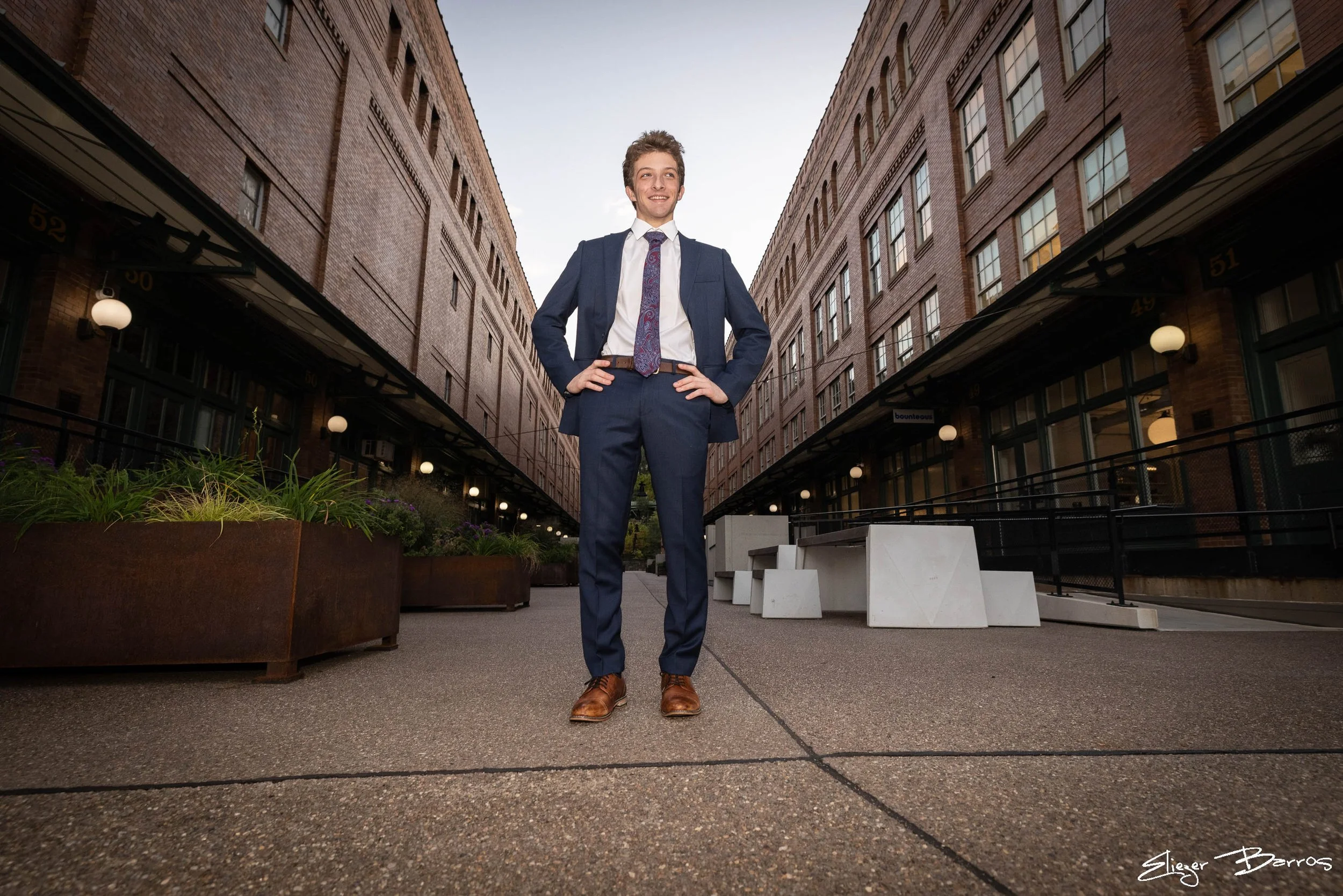A young man in a blue suit with a tie and brown shoes standing confidently in a city alleyway with brick buildings on both sides and a clear sky overhead.