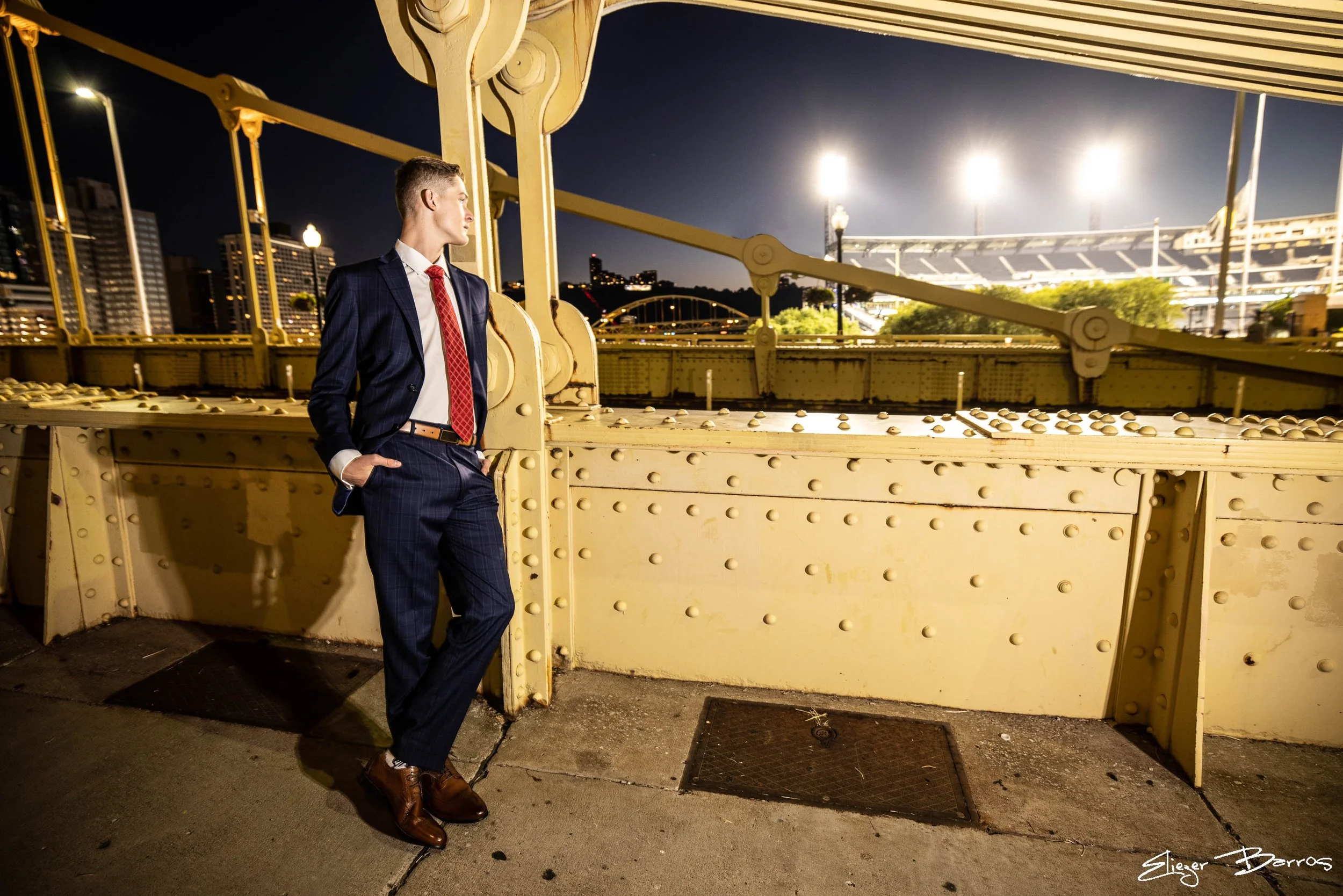 A young man in a dark blue plaid suit, white shirt, red tie, and brown shoes stands on a bridge at night, looking to the side. The bridge has yellow beams and is lit by streetlights, with a city skyline and stadium in the background.