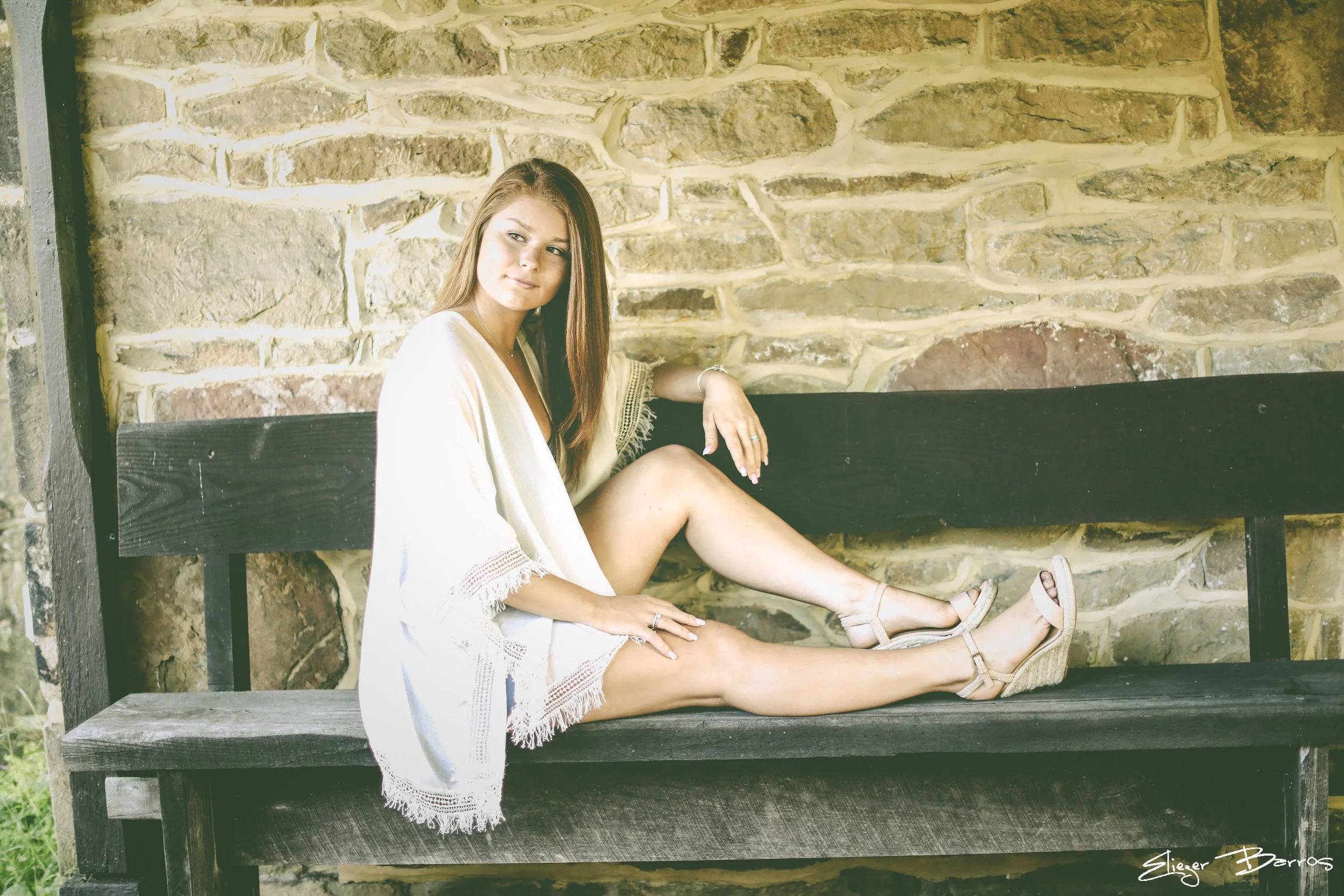 Senior Pictures Pittsburgh at Oliver Miller Homestead. Young woman in a white dress sitting on a wooden bench against a historic stone wall for relaxed and timeless senior portraits in South Park.