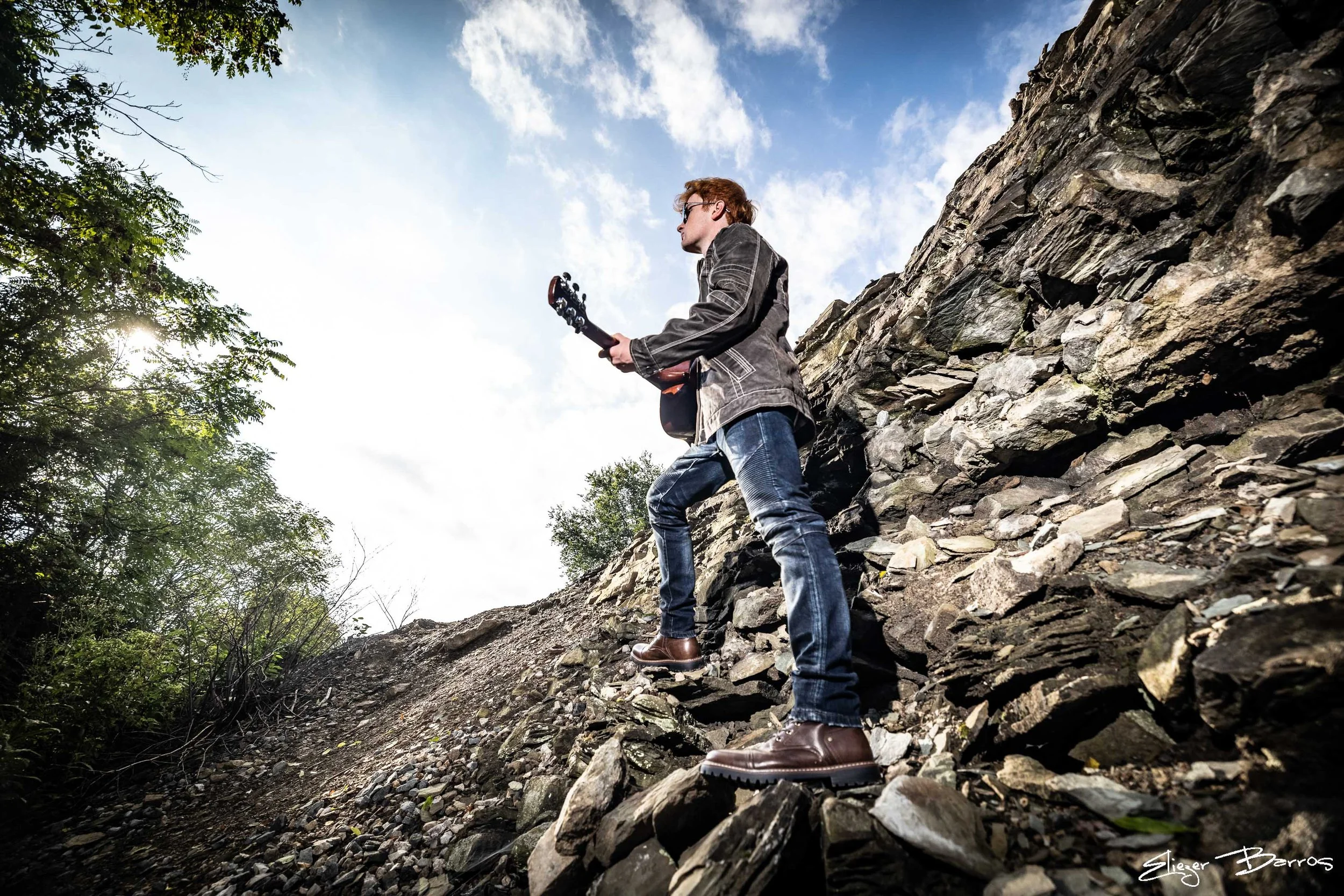 A man with red hair wearing a leather jacket and jeans holding a guitar stands on a rocky slope outdoors with a blue sky and clouds overhead.