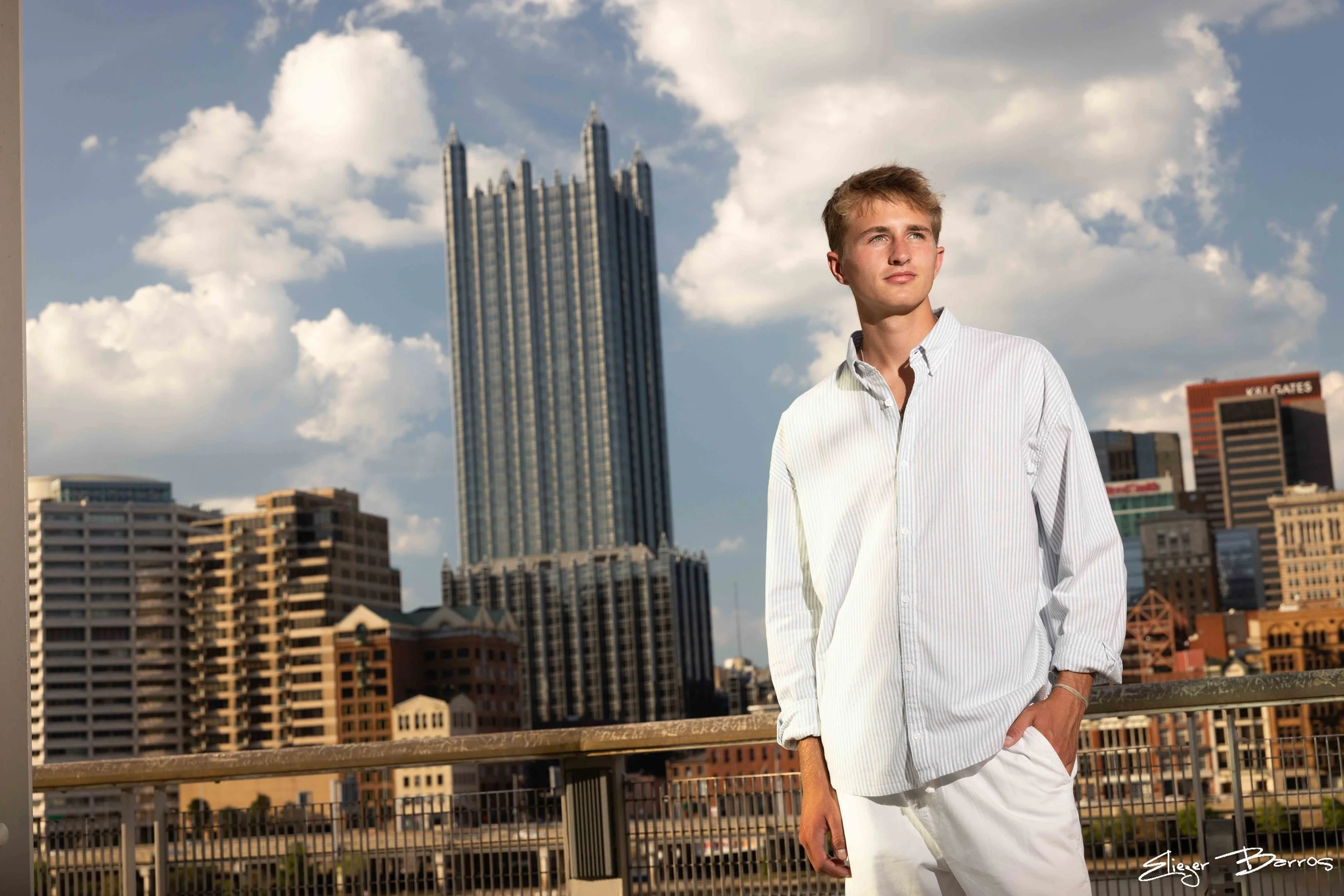 A young man with blond hair wearing a white striped button-up shirt and white pants, standing outdoors on a rooftop with a cityscape and skyscrapers in the background during daytime.
