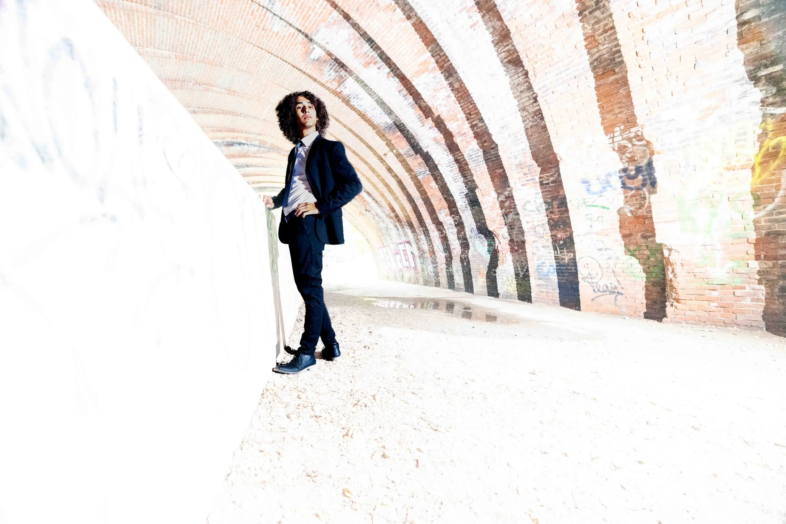 A young man with curly hair dressed in a black suit, white shirt, and black boots, standing in under a brick tunnel with graffiti on the wall, looking confidently at the camera.