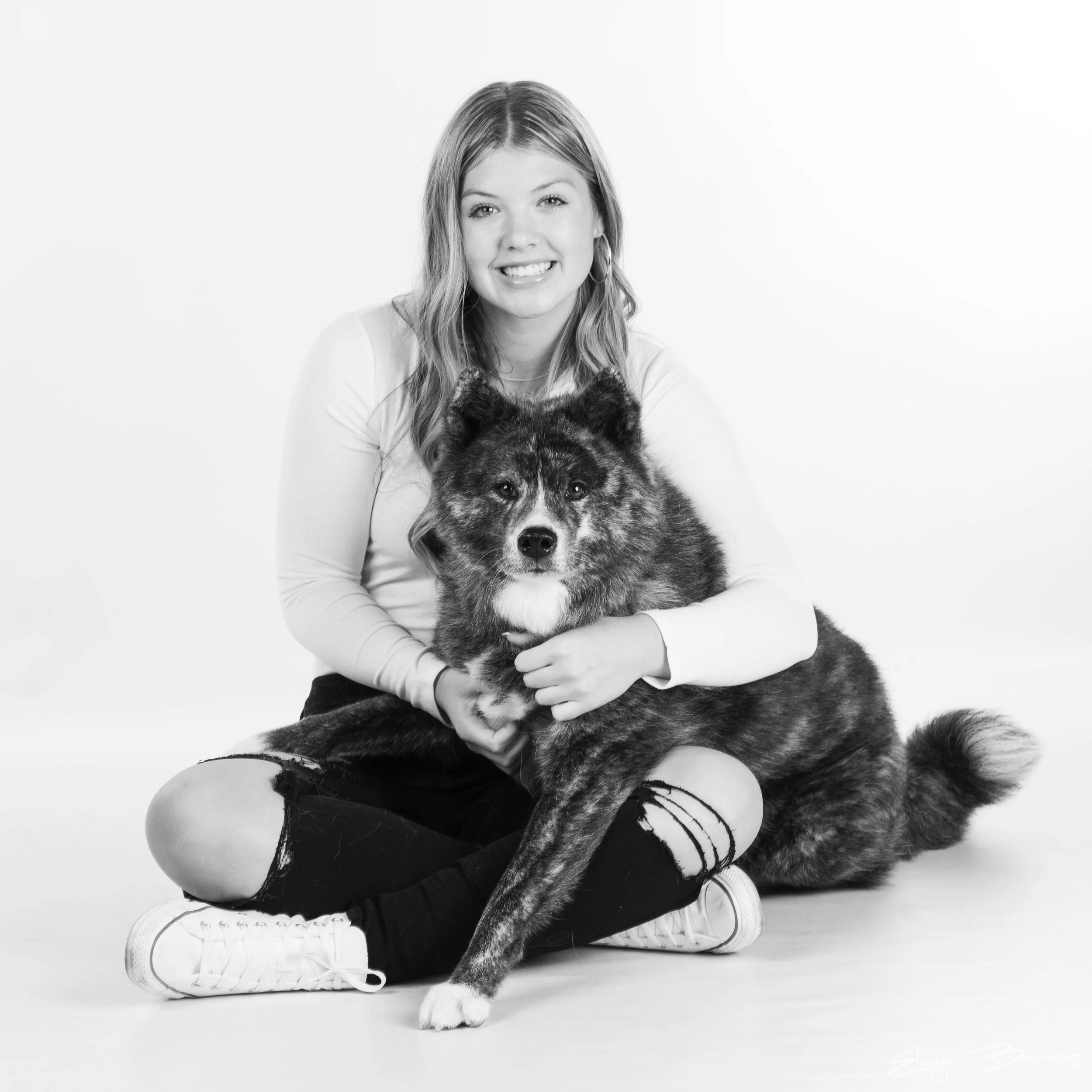 Black and white photo of a young woman sitting cross-legged on a light-colored floor, holding a large, fluffy, and calm dog. The woman is smiling, has long hair, and is wearing casual clothing including a long-sleeve shirt and sneakers. The backgroun