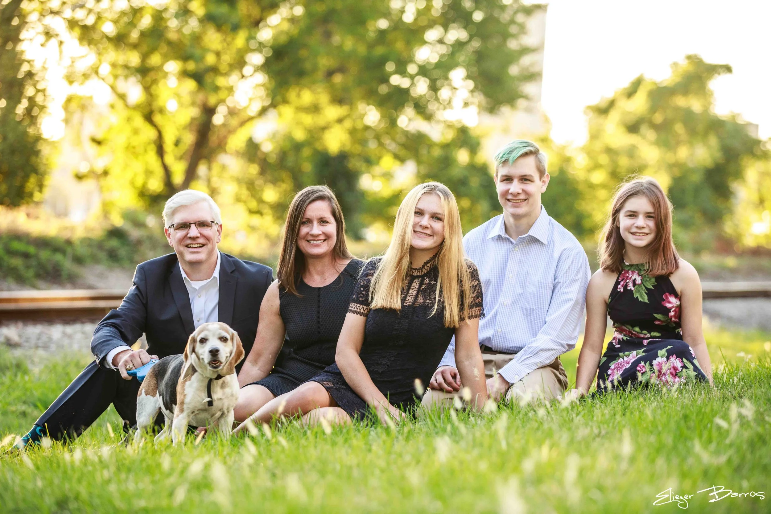 A multigenerational family group sitting on grass outdoors with trees in the background, including three women, two young men, an older man, and a dog.