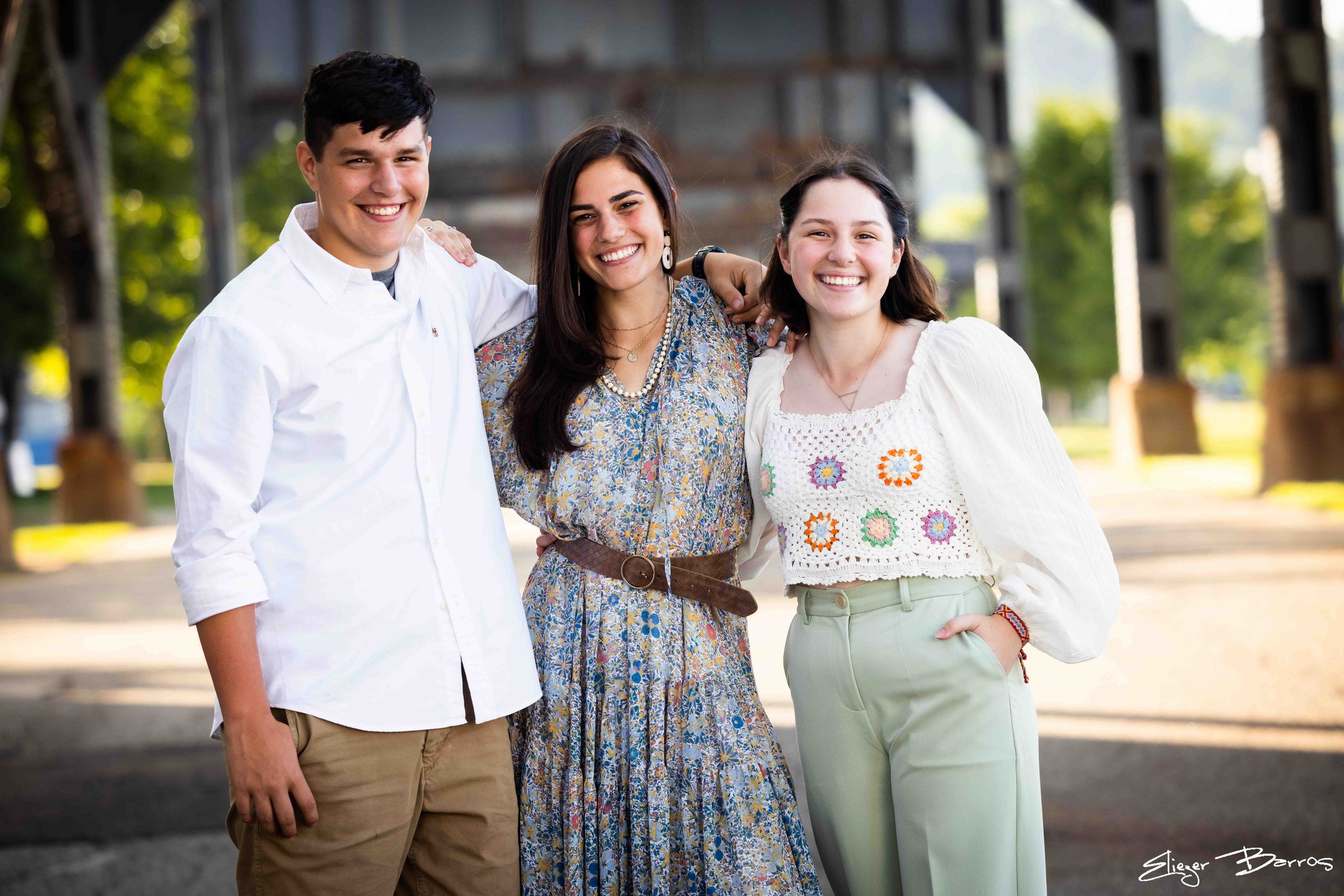 Three young adults, two women and one man, smiling and standing close together outdoors with trees and an industrial structure in the background.
