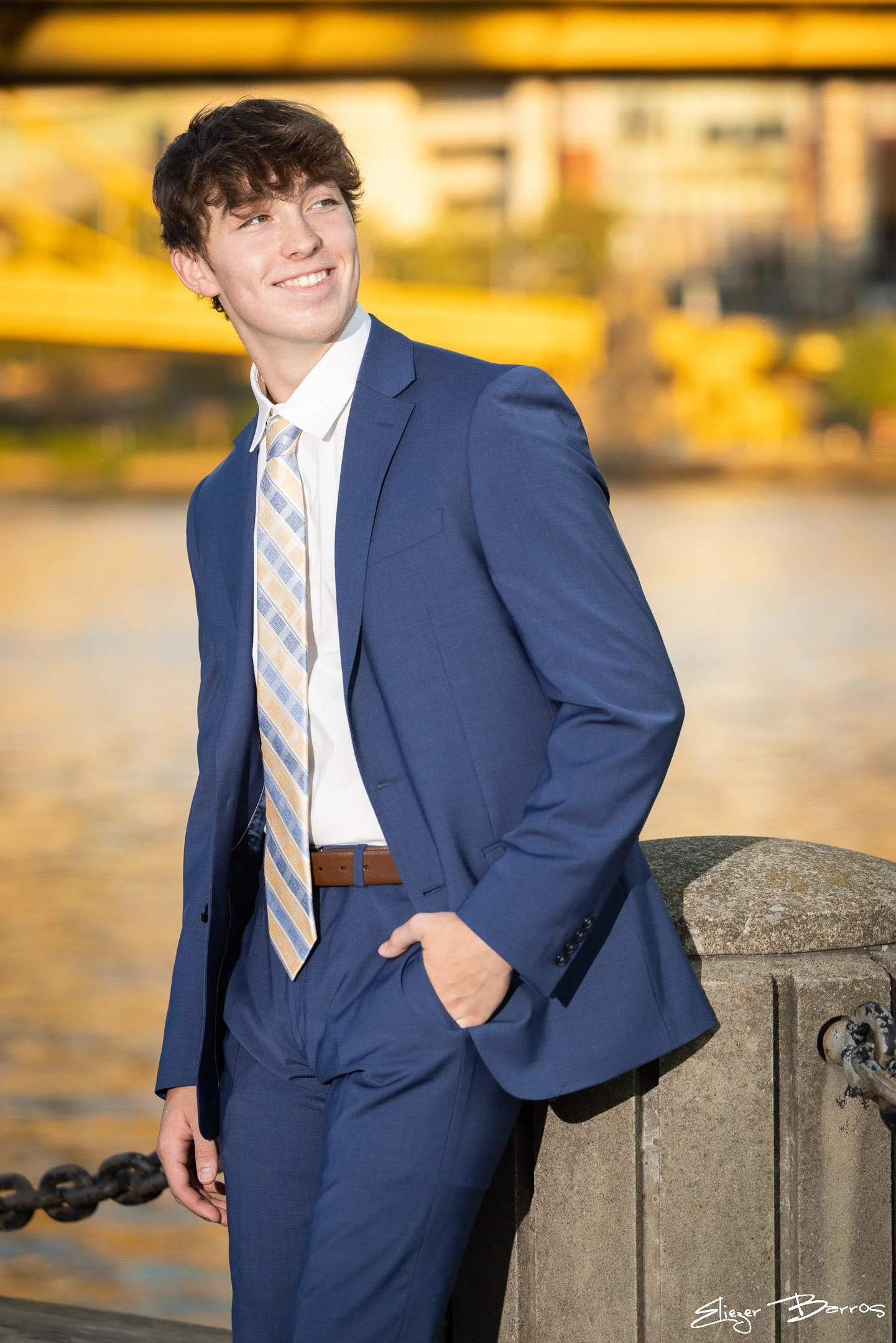 A young man in a blue suit and tie leans against a stone railing by a body of water during sunset. He has light brown, curly hair and is smiling while looking to his left.