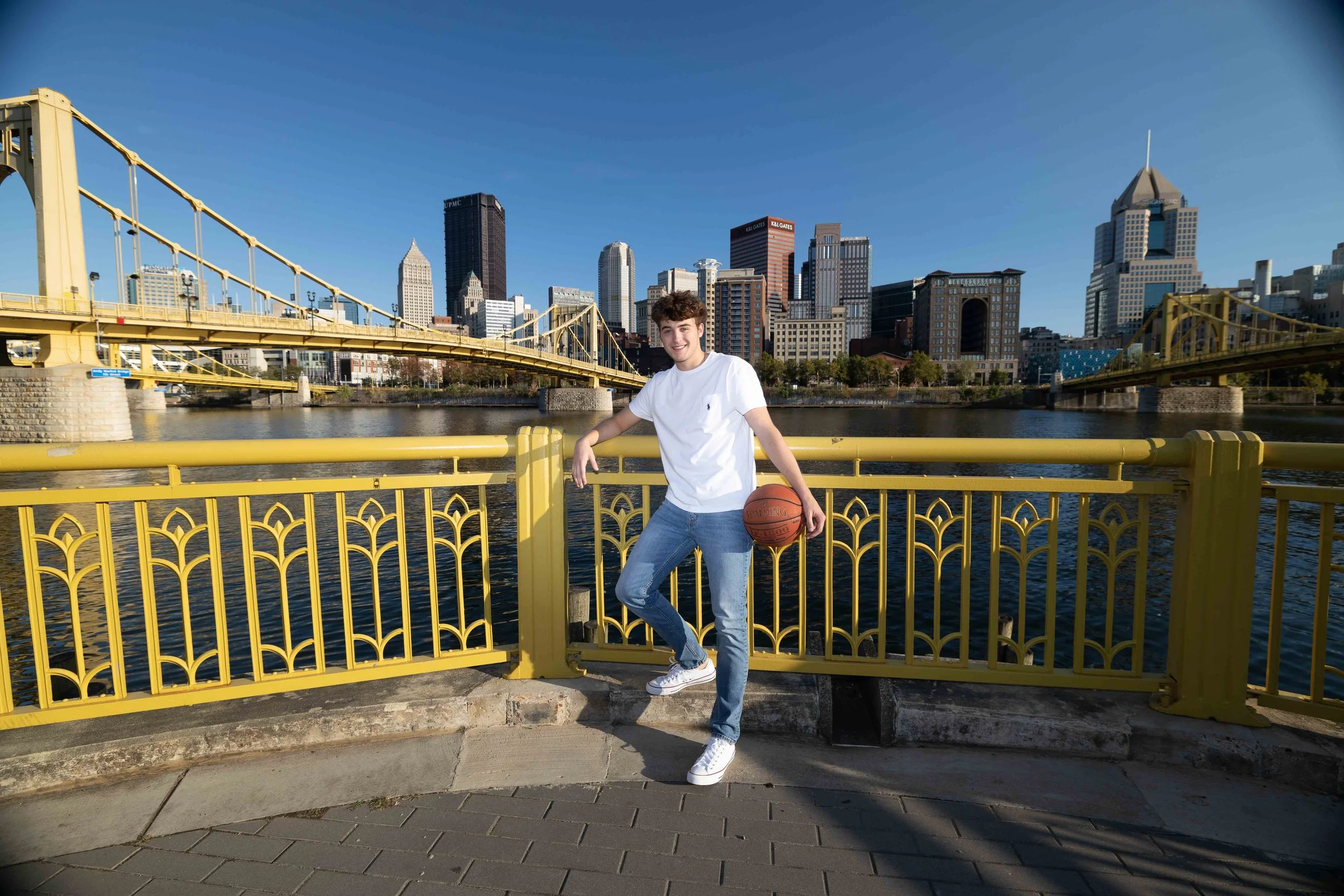 A young man standing by a yellow railing on a bridge, holding a basketball, with a city skyline and a river in the background on a clear sunny day.