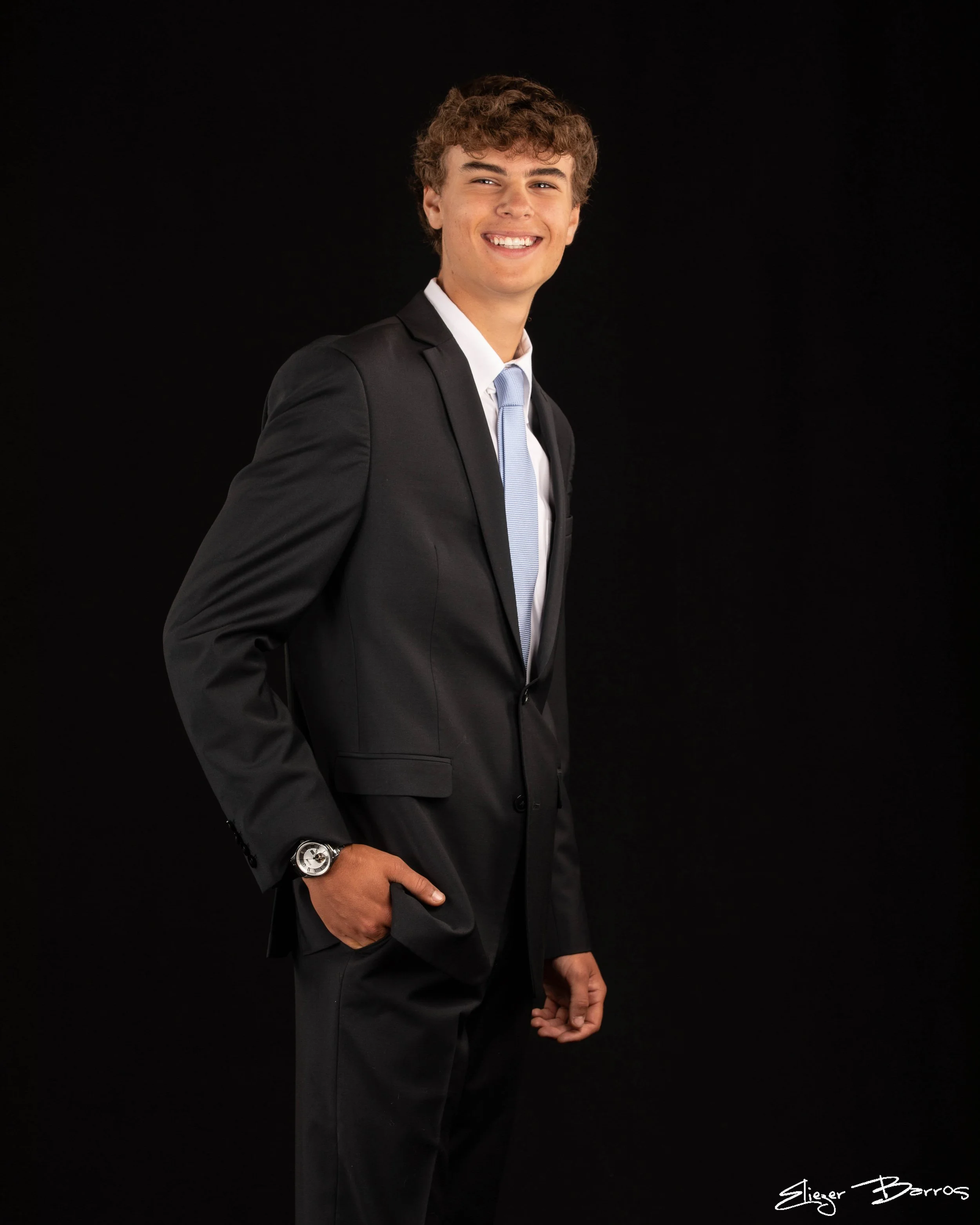 Young man in a suit smiling, standing against a black background, with hand in pocket and wearing a wristwatch.