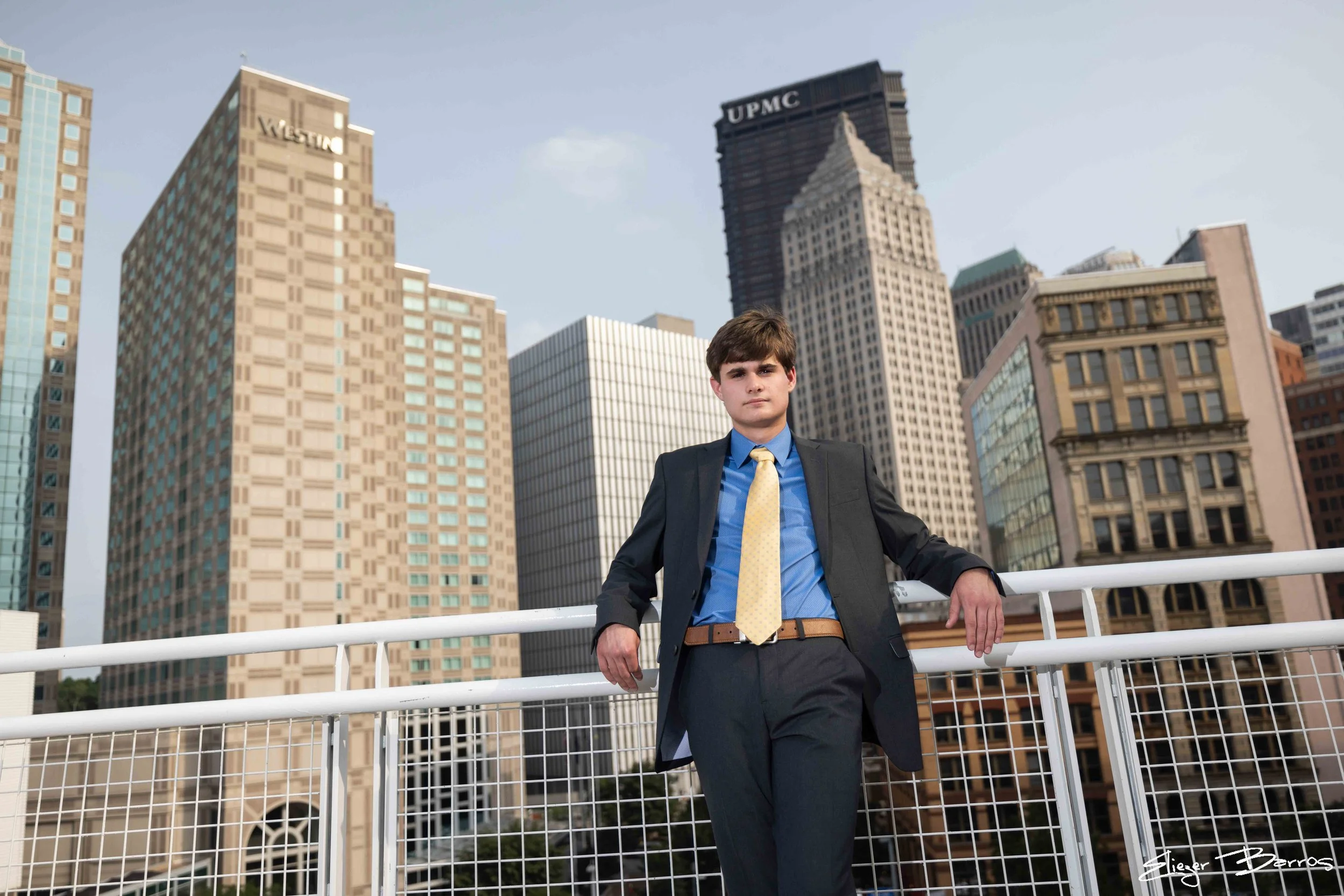 Young man in a formal suit standing with arms resting on a white railing, cityscape background with tall buildings, some branded with Westin and UPMC logos, under a clear sky.
