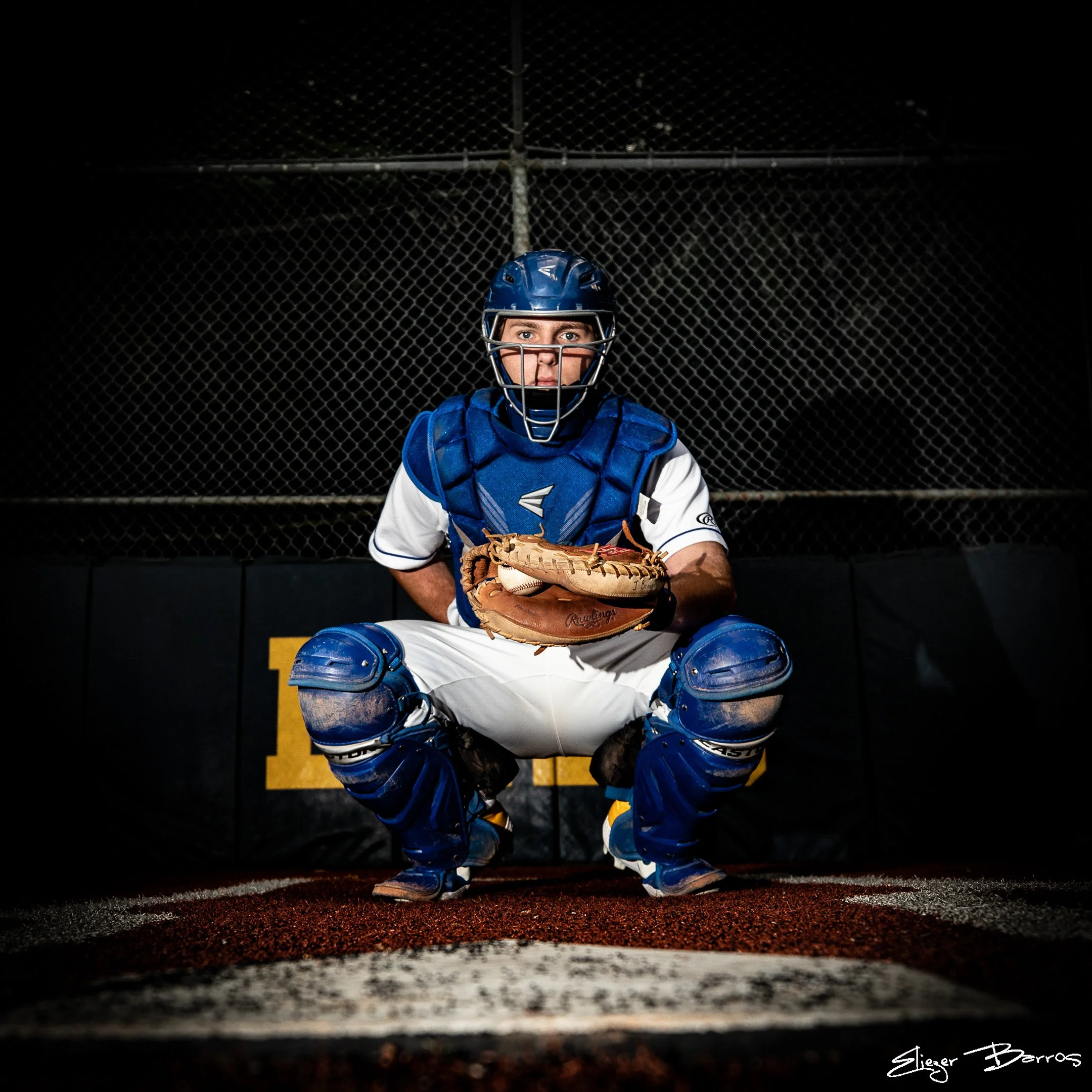 A young baseball player dressed in catcher's gear squatting behind home plate, holding a baseball glove, with a chain-link fence and dark night sky in the background.