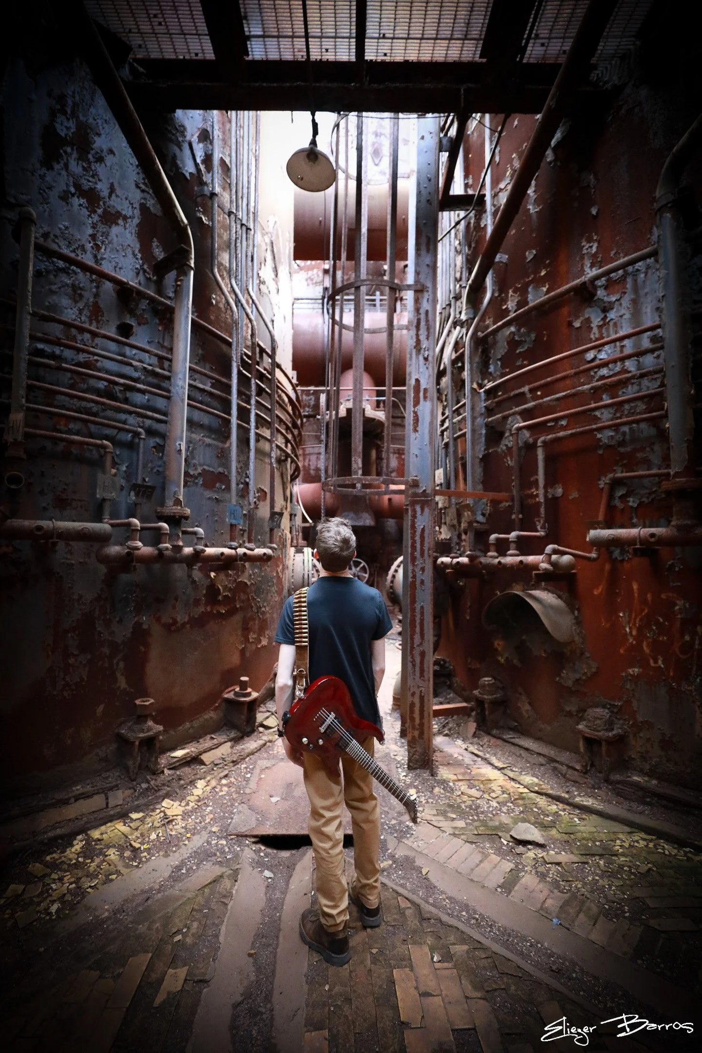A person holding a red electric guitar walks through an abandoned, rusty industrial space with pipes and peeling walls, looking up at the ceiling.