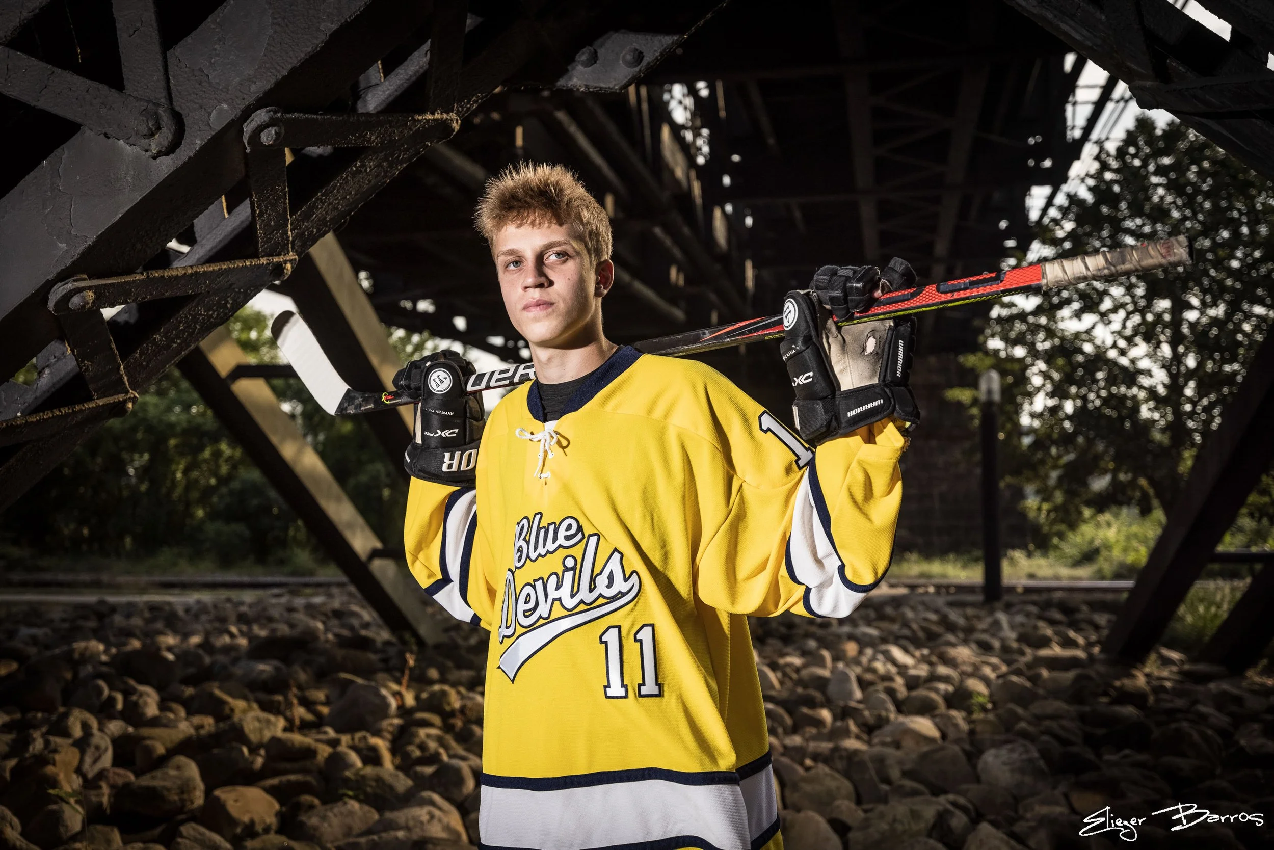 A young man in a yellow hockey jersey holding a hockey stick over his shoulder, standing under a bridge with trees in the background.