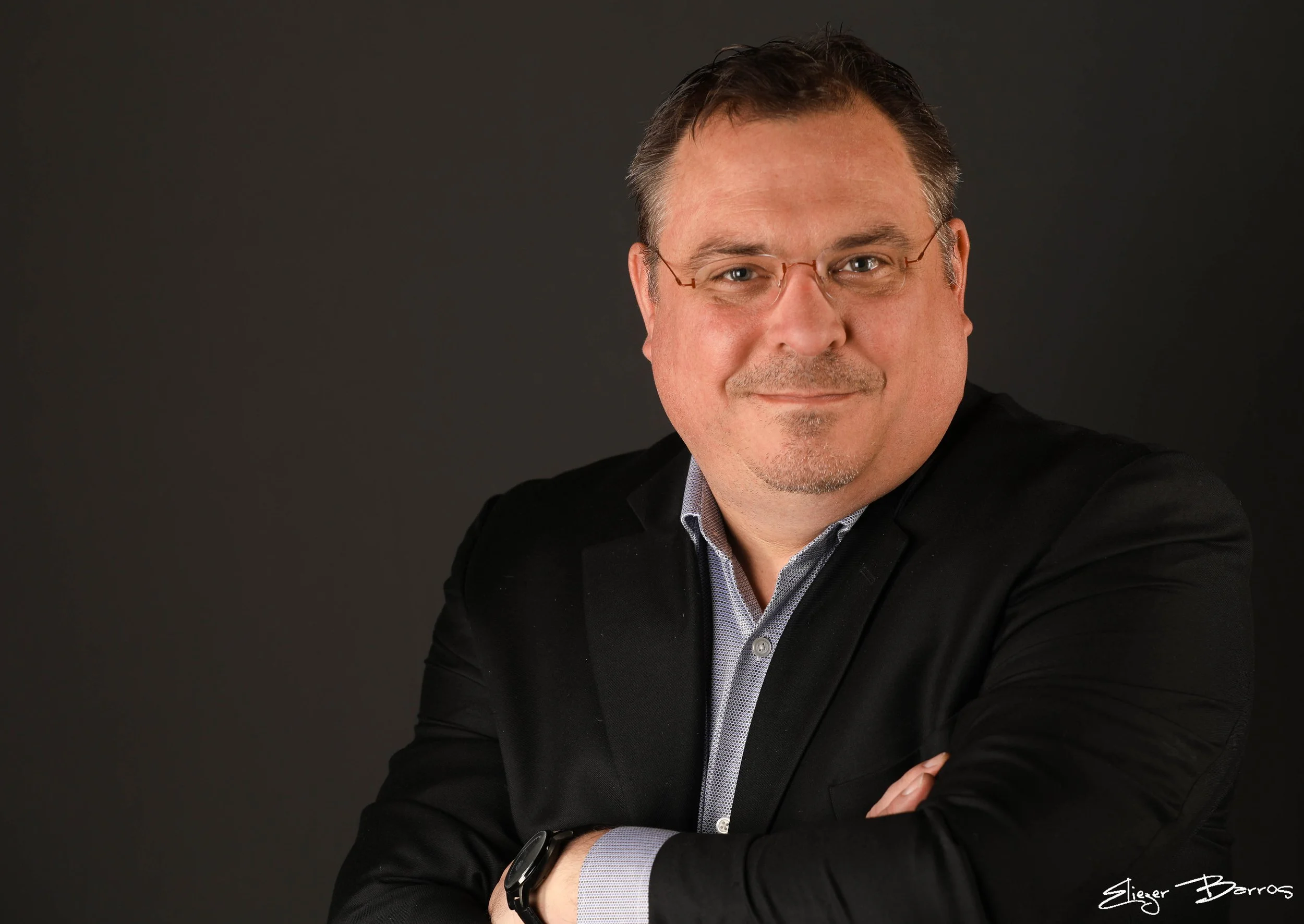 A man with glasses and short dark hair, wearing a black blazer and patterned shirt, smiling against a dark background.