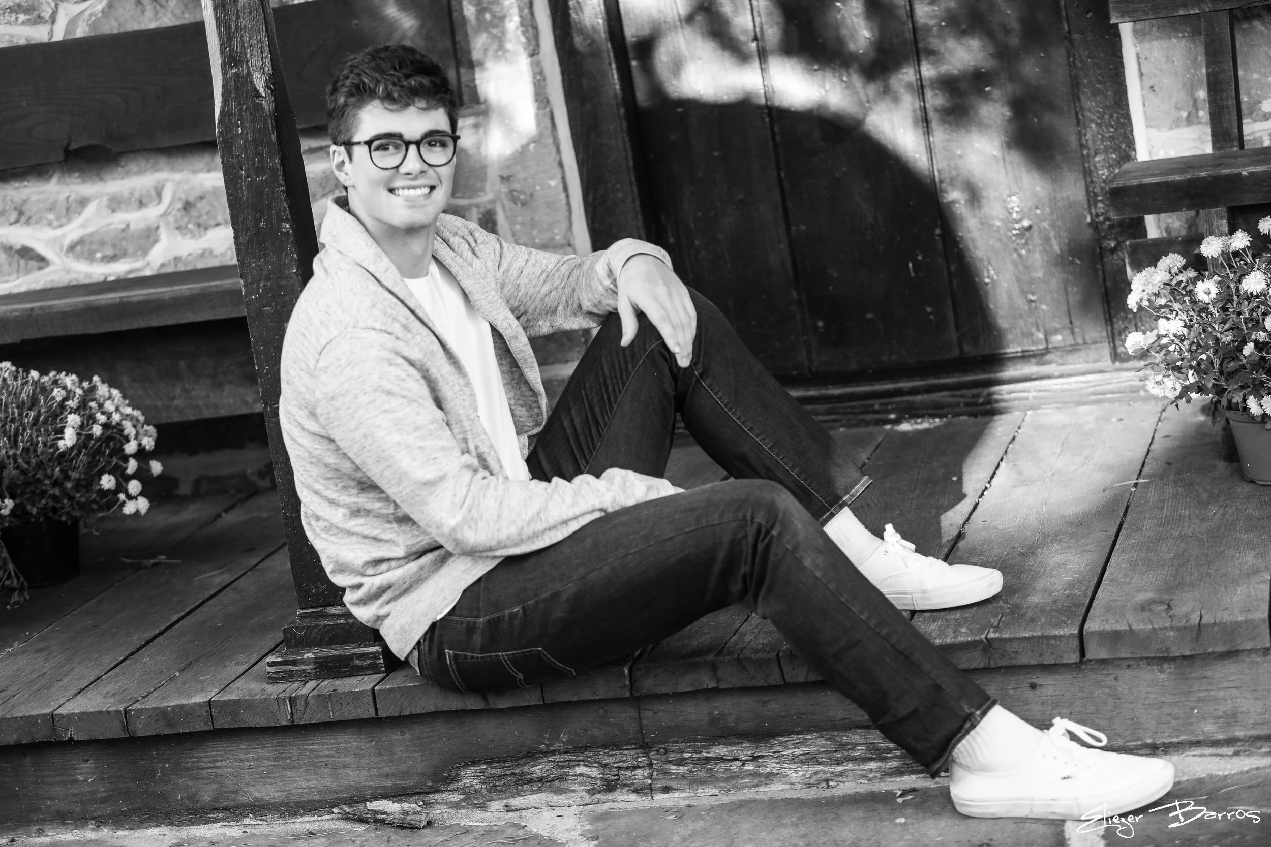 A young man with glasses, wearing a light-colored hoodie, white shirt, jeans, and white sneakers, sits on a wooden porch with potted flowers nearby, smiling at the camera.