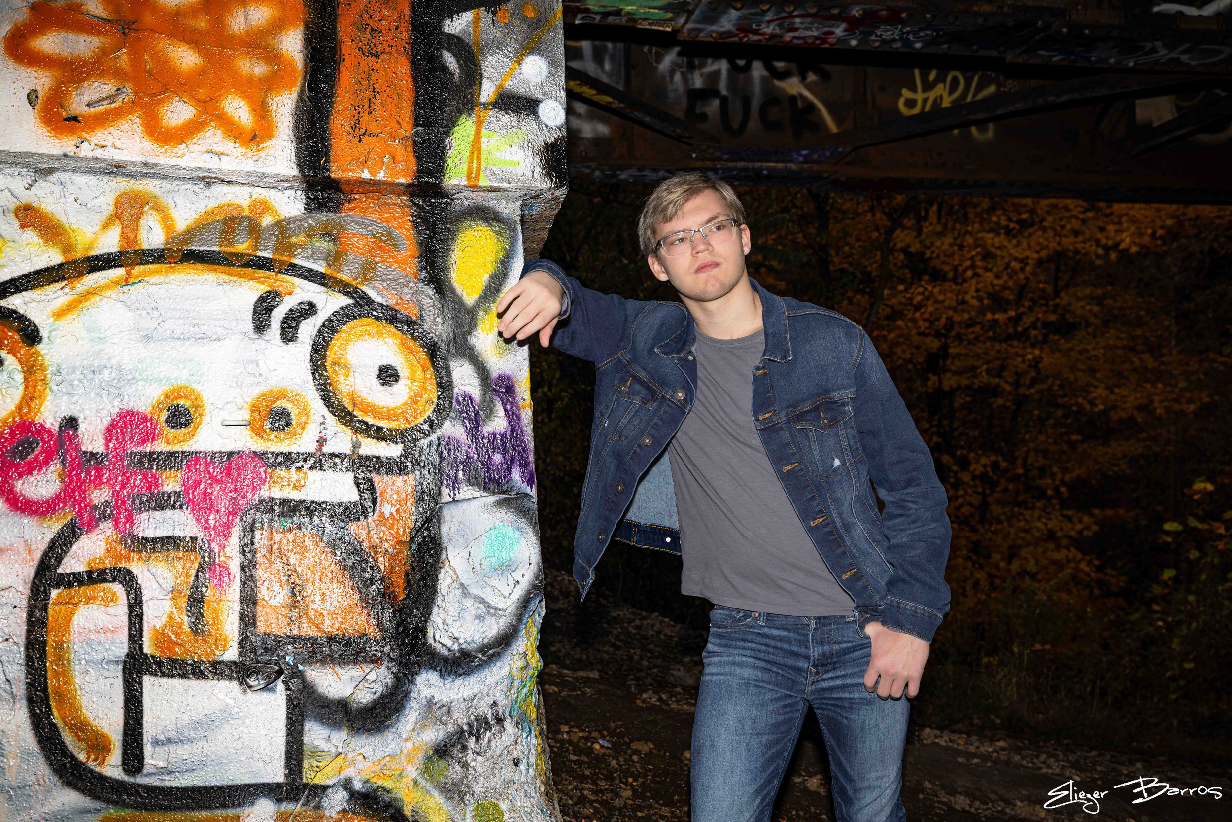 A young man in glasses and a denim jacket standing next to a graffiti-covered wall in an outdoor setting at night, with trees in the background.