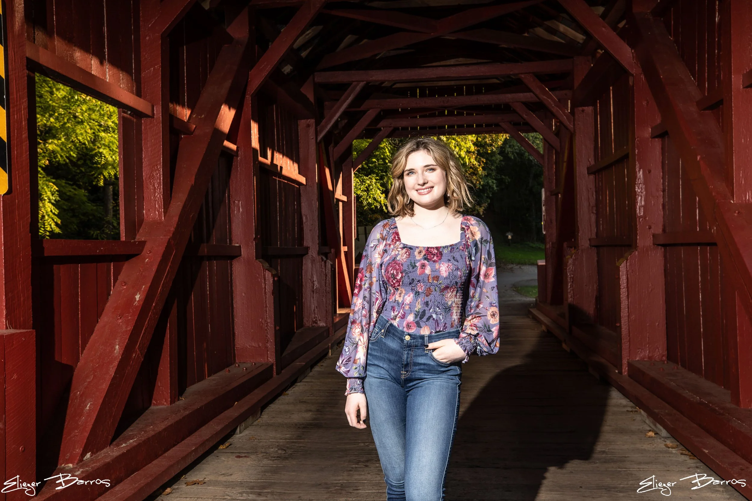 A young woman with shoulder-length wavy hair wearing a floral blouse and jeans, smiling as she stands under a red wooden bridge with greenery in the background.