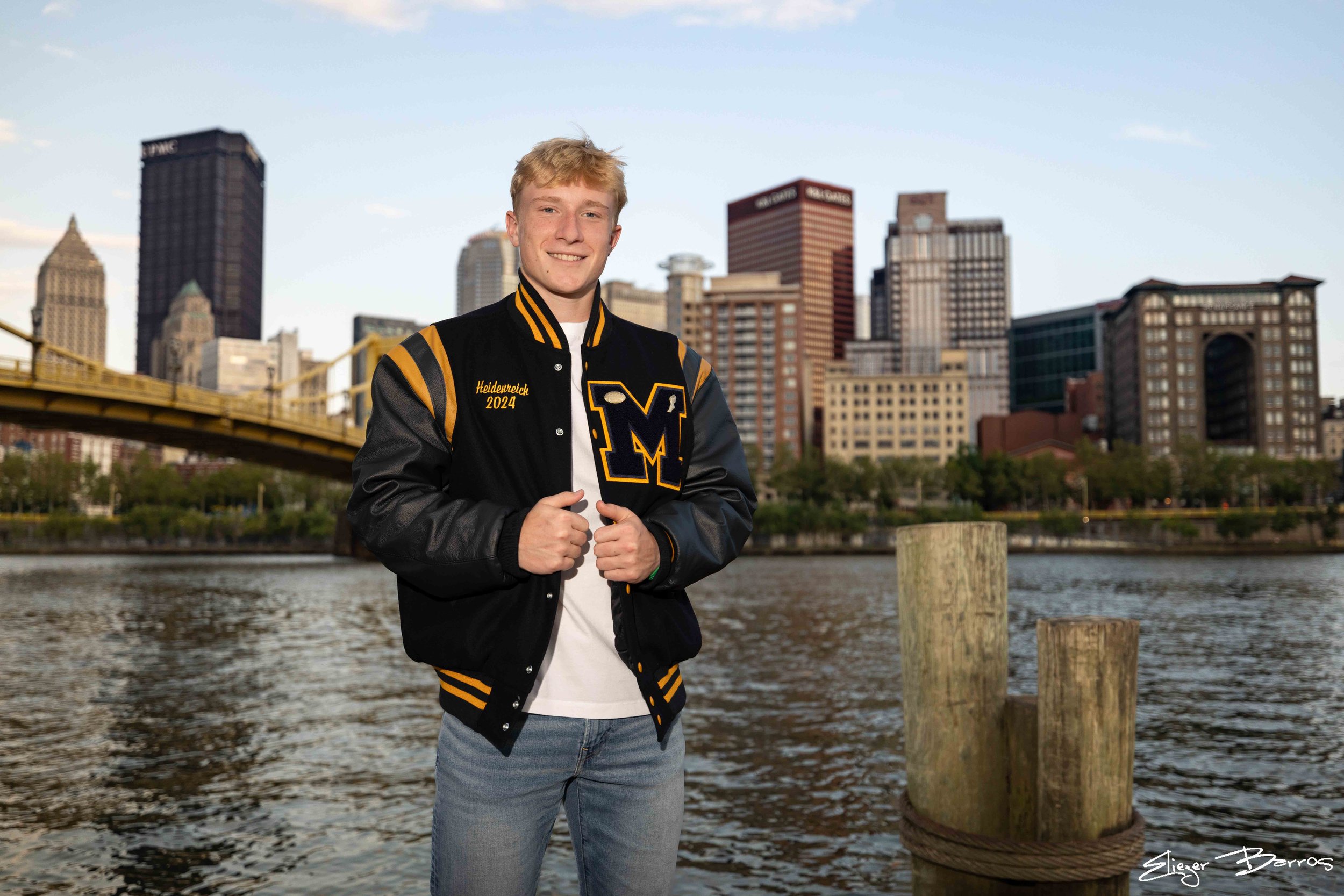A young man with blond hair smiling and standing by a river in a city with tall buildings and a yellow bridge in the background, wearing a black varsity jacket with yellow accents and the name 'Heidenreich 2024' on it.