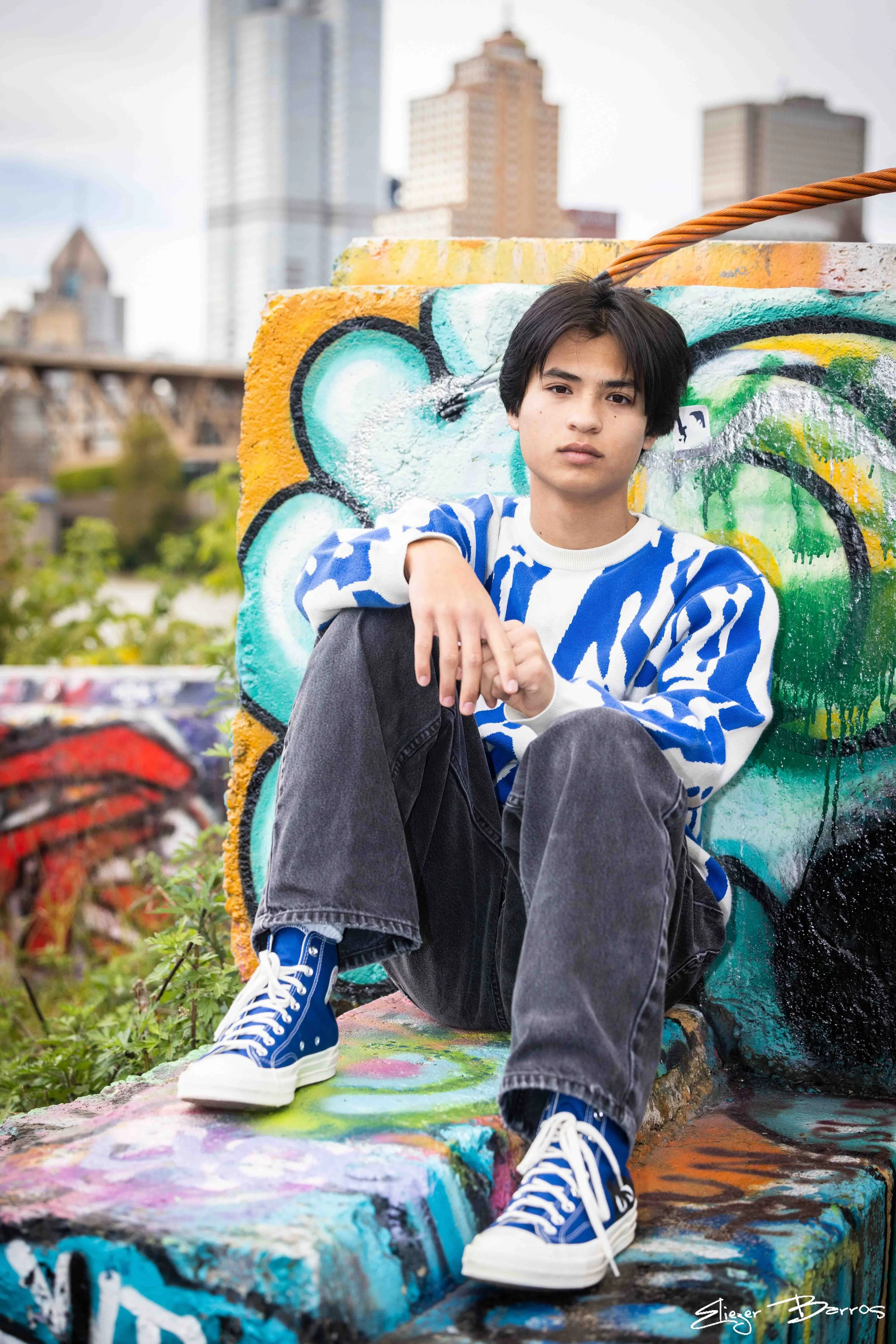A young man with black hair sitting on a colorful graffitied concrete ledge in an urban park setting with city buildings in the background. He's wearing a blue and white patterned sweater, black jeans, and blue high-top sneakers. The sky is partly cl