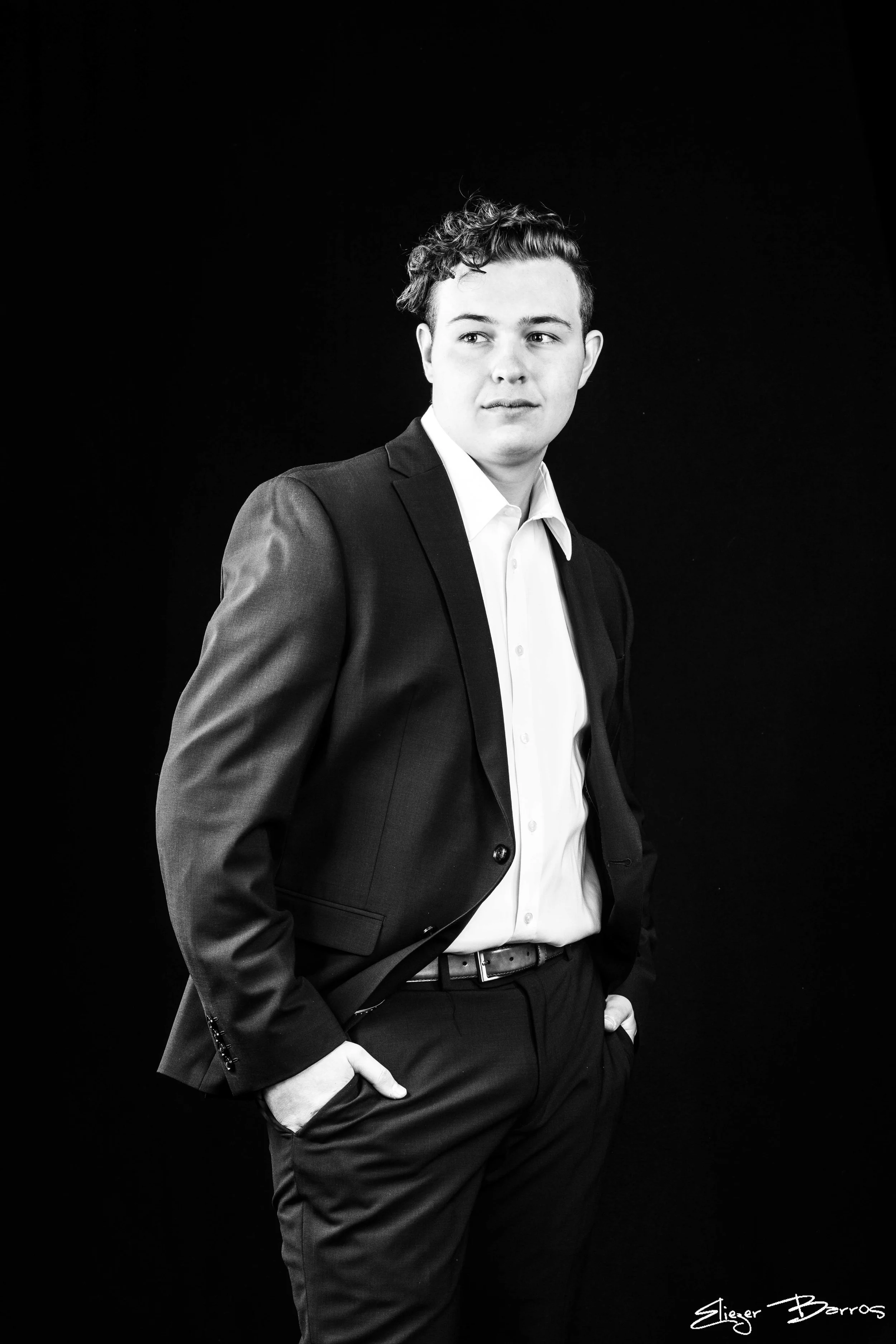 A young man in a suit with one hand in his pocket, standing against a black background, looking slightly to the side. Black and white photo.