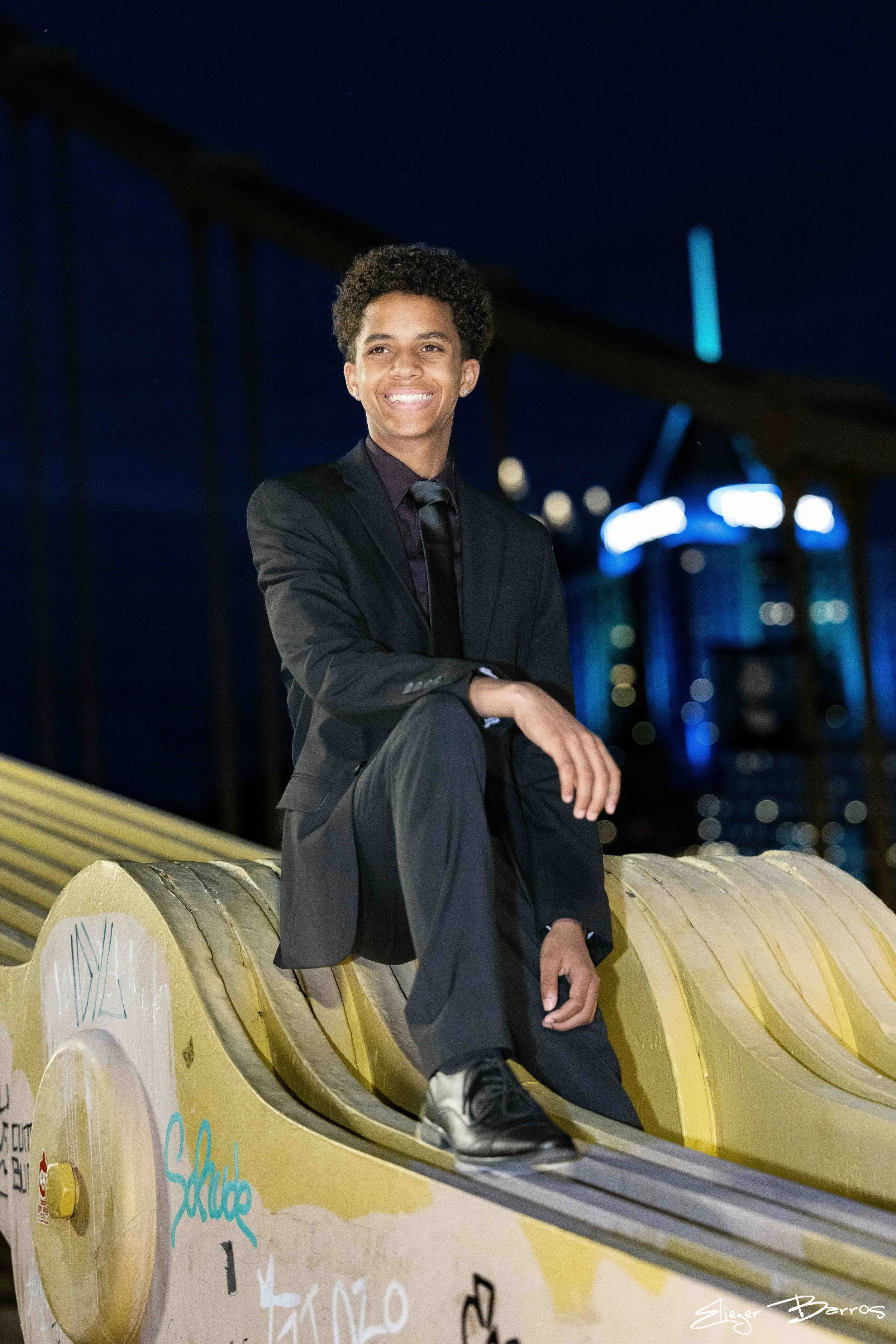 A young man in a black suit sitting on a yellow bench at night, smiling with his arms crossed, with a bridge and city lights in the background.