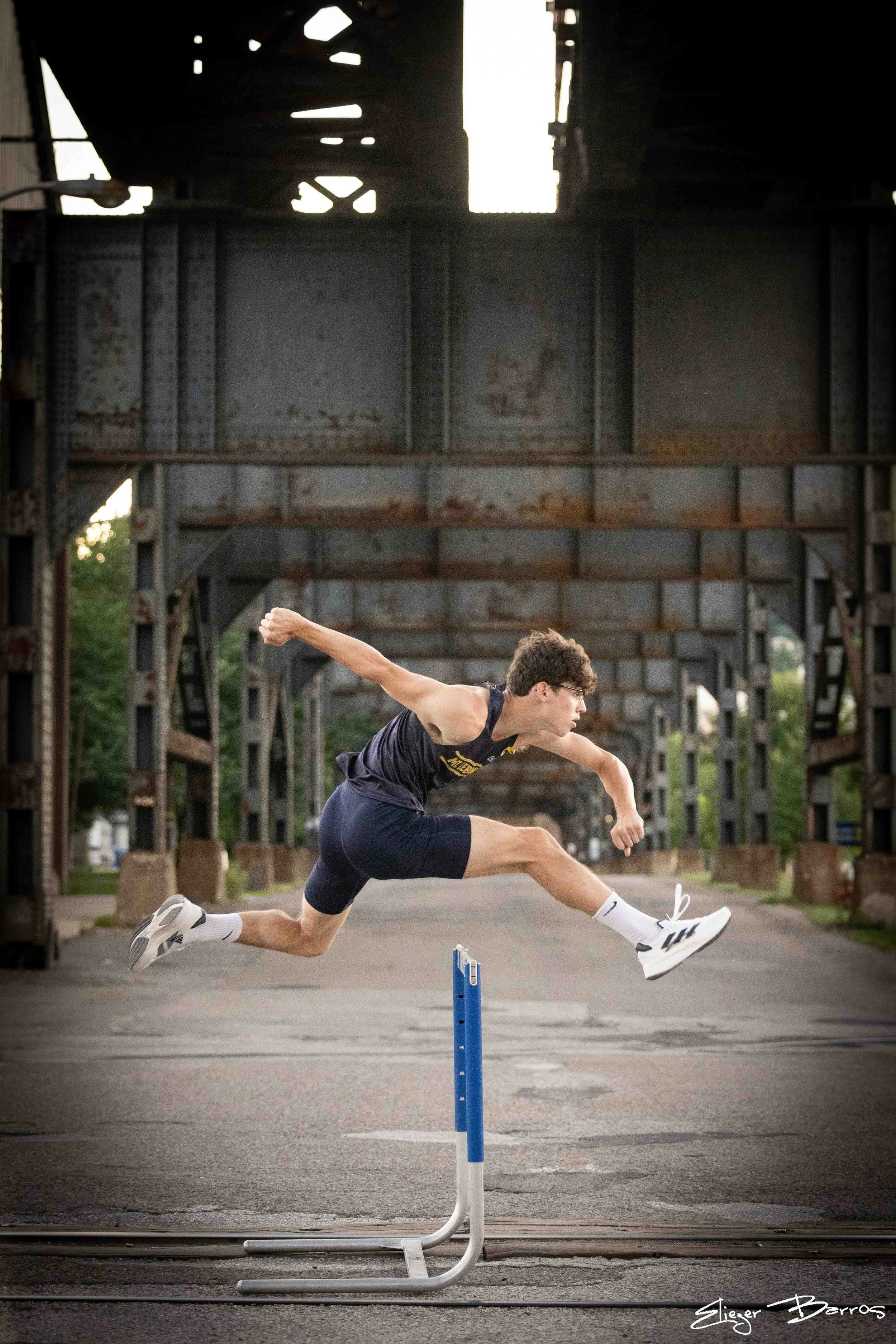A male athlete in sportswear jumping over hurdles on an outdoor track during daytime under an overpass.