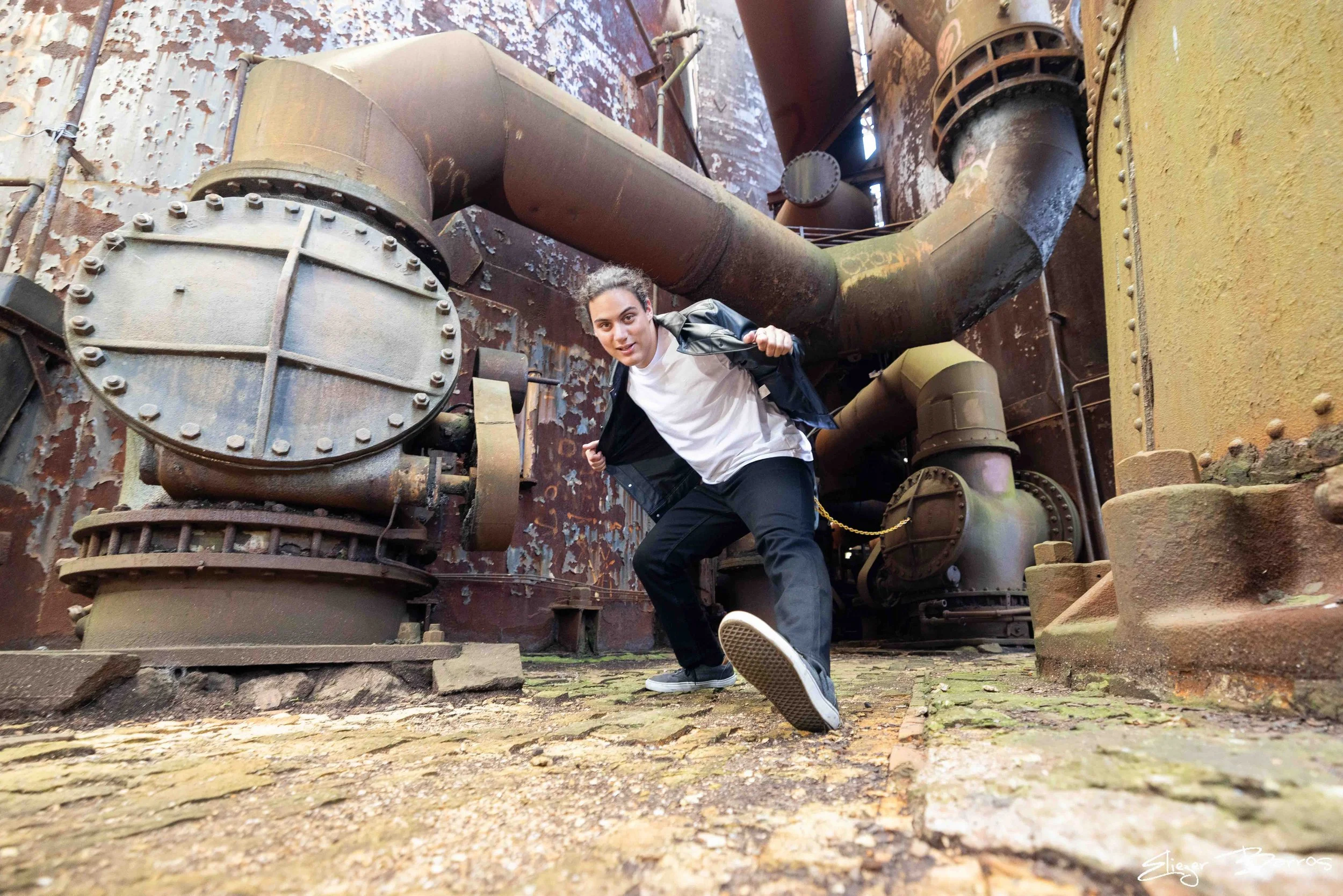 Young woman in casual clothing climbing or crouching among large, rusty industrial pipes and machinery inside a weathered, abandoned factory or industrial building.