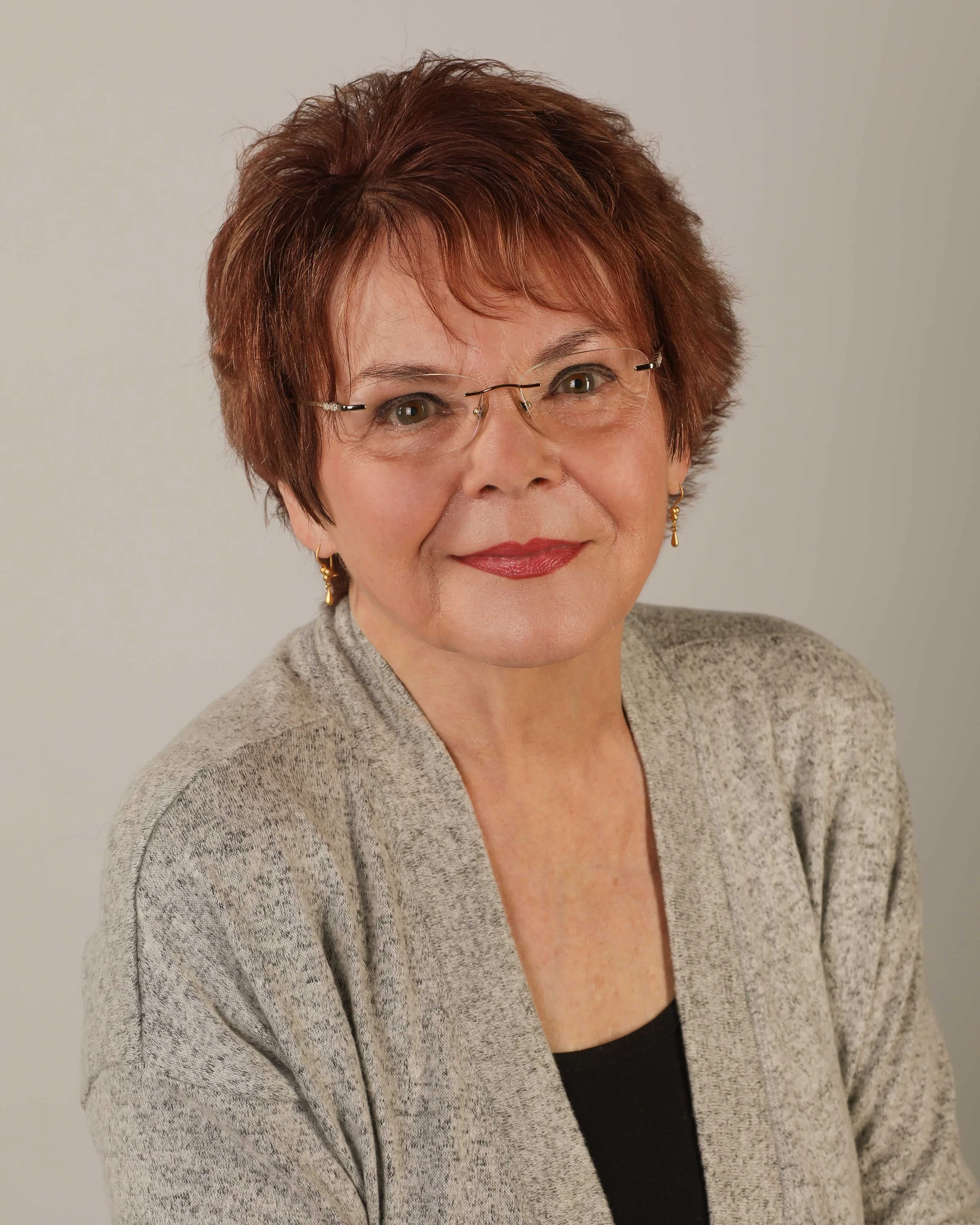 Close-up of a middle-aged woman with short, reddish-brown hair, wearing glasses, red lipstick, gold earrings, a gray sweater, and a black top, against a plain light background.