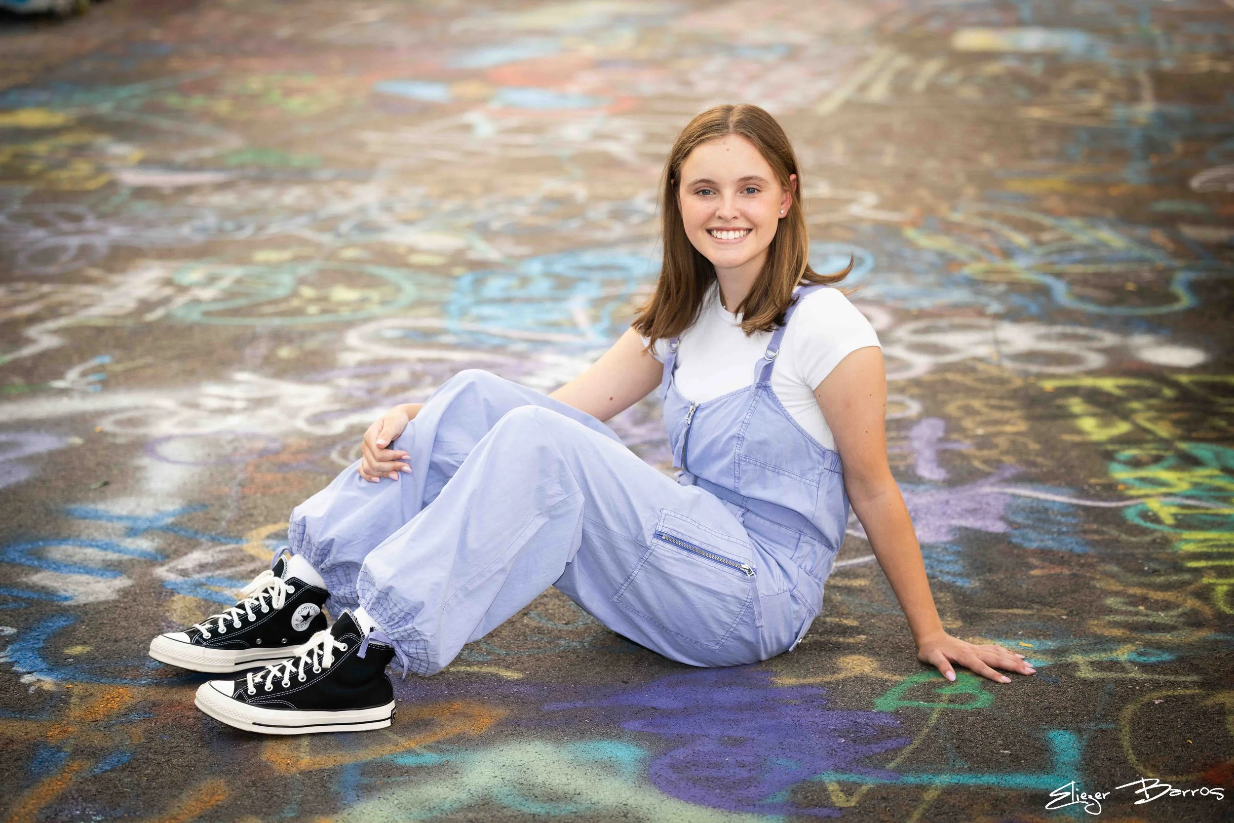 A young woman with brown hair and a white t-shirt, wearing a light purple jumpsuit, stylish black and white high-top sneakers, sits on colorful chalk drawings on a concrete sidewalk, smiling at the camera.