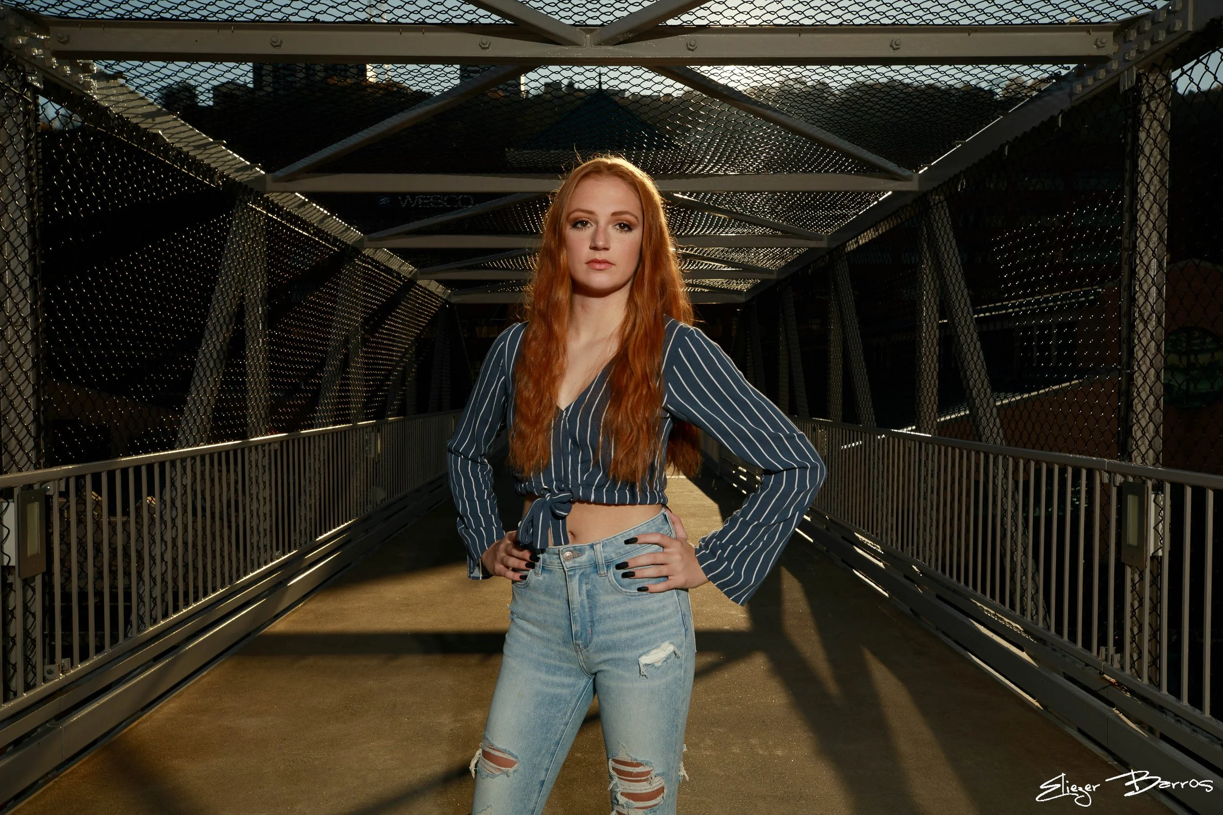 Red-haired woman in striped shirt and ripped jeans standing on a pedestrian bridge with metal railings, sunset sky in the background.