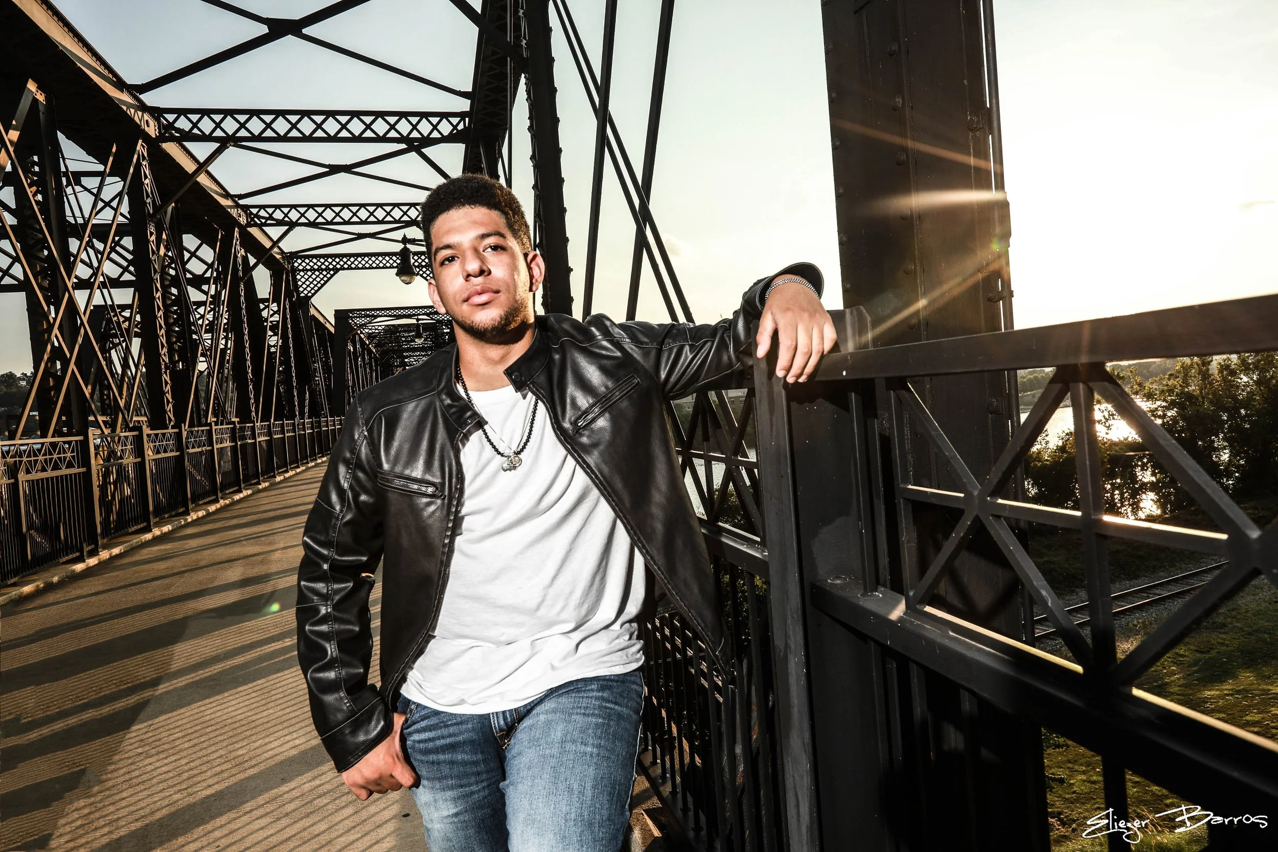 A young man in a black leather jacket and white t-shirt standing on a bridge, leaning on the railing, with a sunset sky and trees in the background.