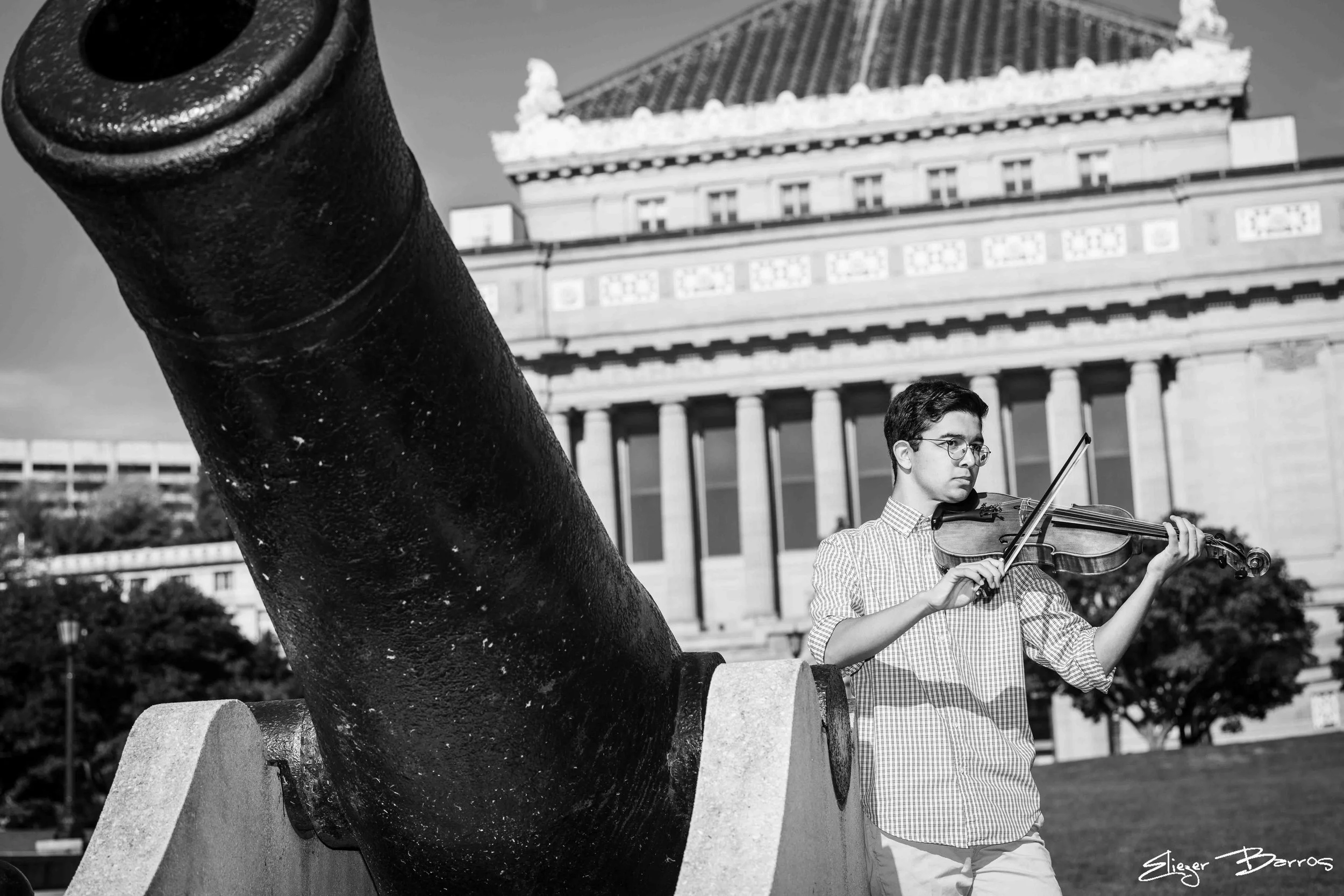 A young man playing the violin outdoors near a large statue and a historic building with columns in the background.