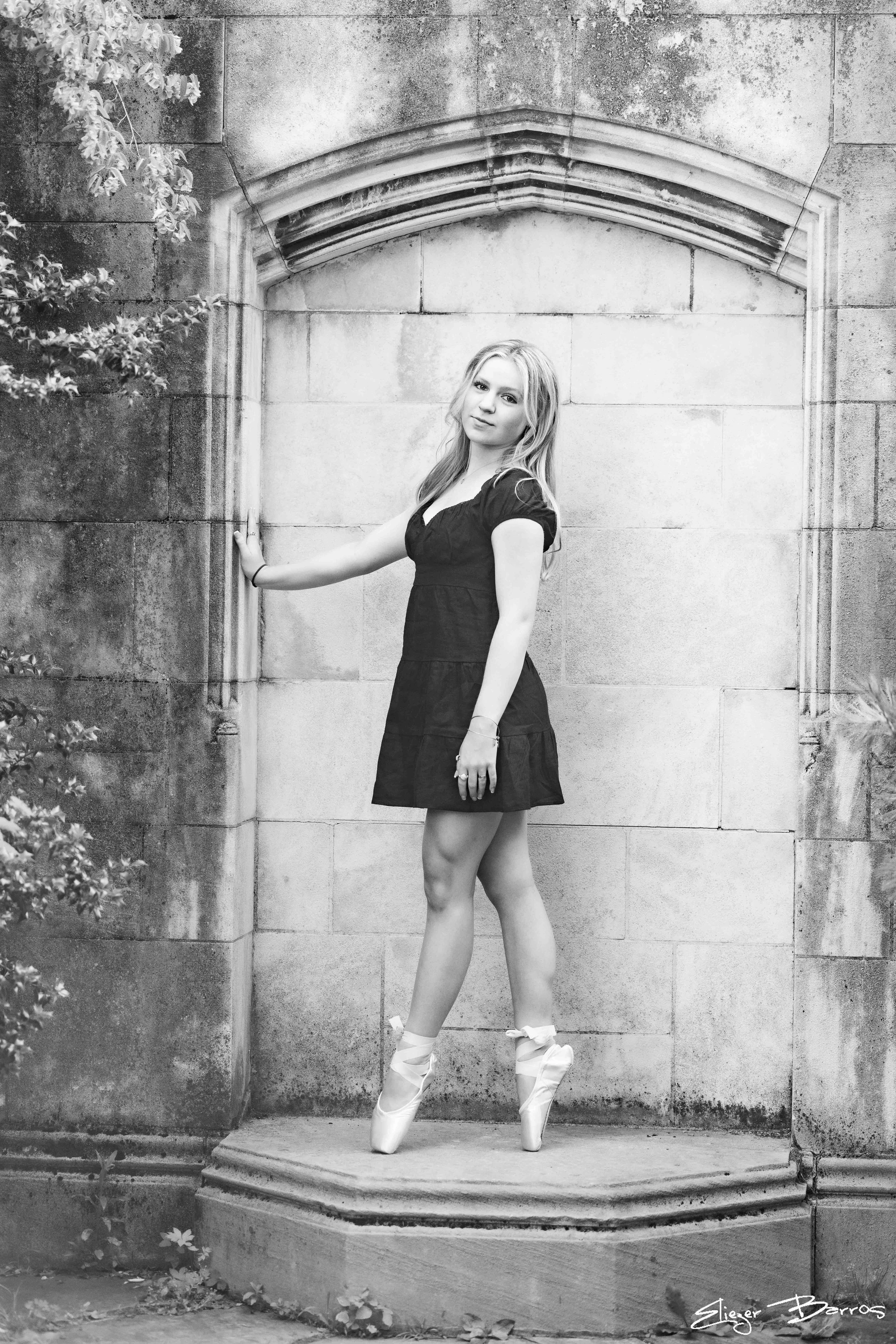 Black and white photo of a young woman standing on pointed ballet shoes at the entrance of a stone building, with her arm extended to touch the stone wall. She is wearing a short dress and has loose hair. There is foliage on the left side of the imag