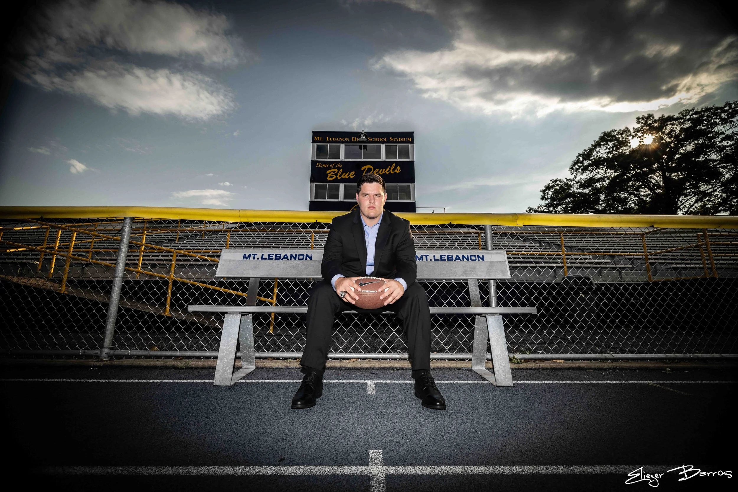 A young man in a black suit sitting on a bench at a football stadium, holding a football. The stadium sign reads "Mt. Lebanon High School Stadium," and the words "Home of the Blue Devils" are on the grandstand. The sky is partly cloudy, and the sun i
